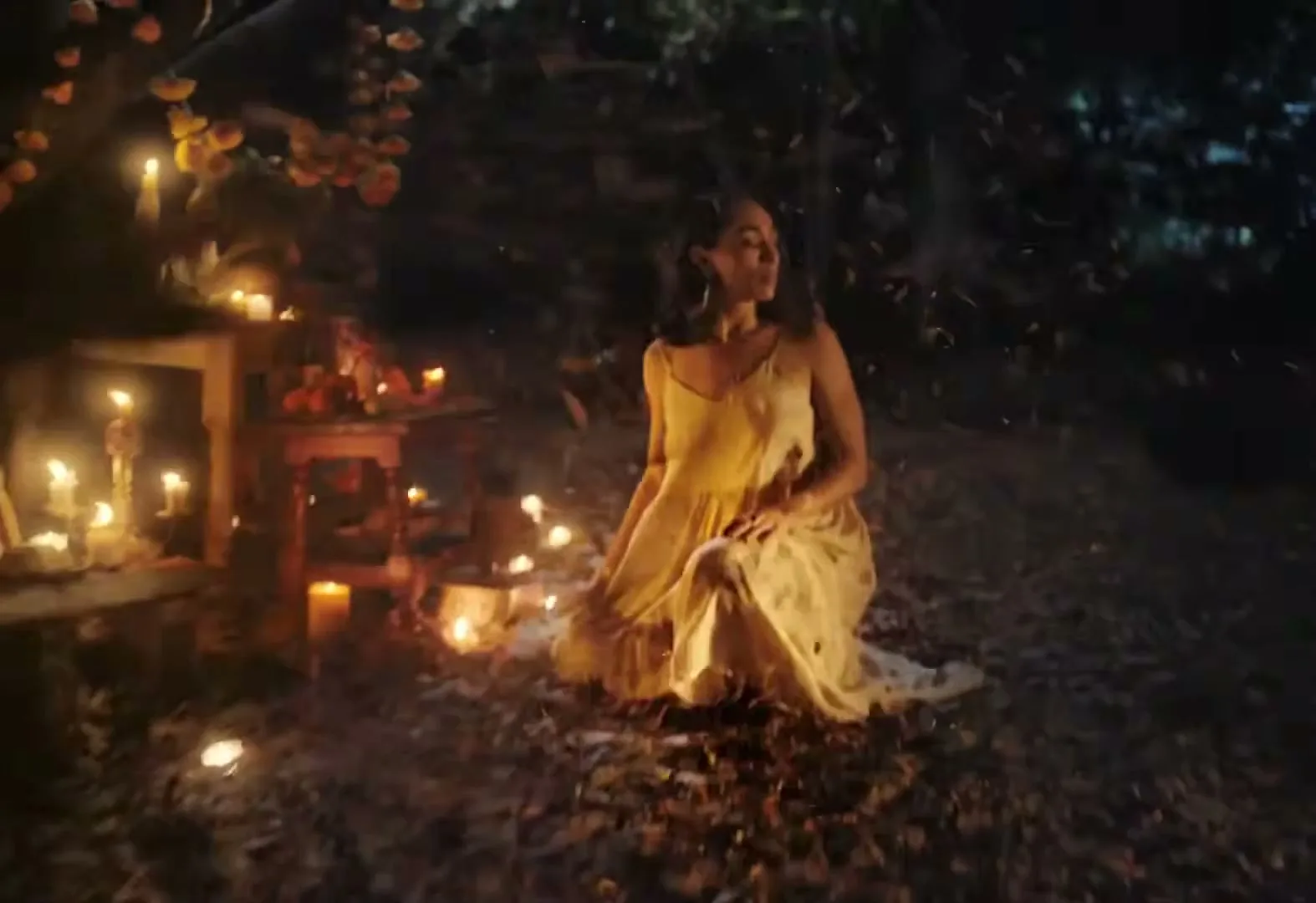 A woman in a light dress kneels on the ground surrounded by lit candles and falling leaves at night, next to an altar.