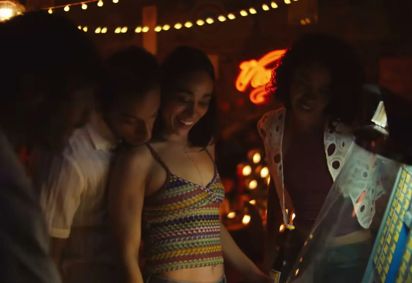 A group of young adults, including a woman in a colorful top, gather around a jukebox in a dimly lit bar, smiling and enjoying themselves.