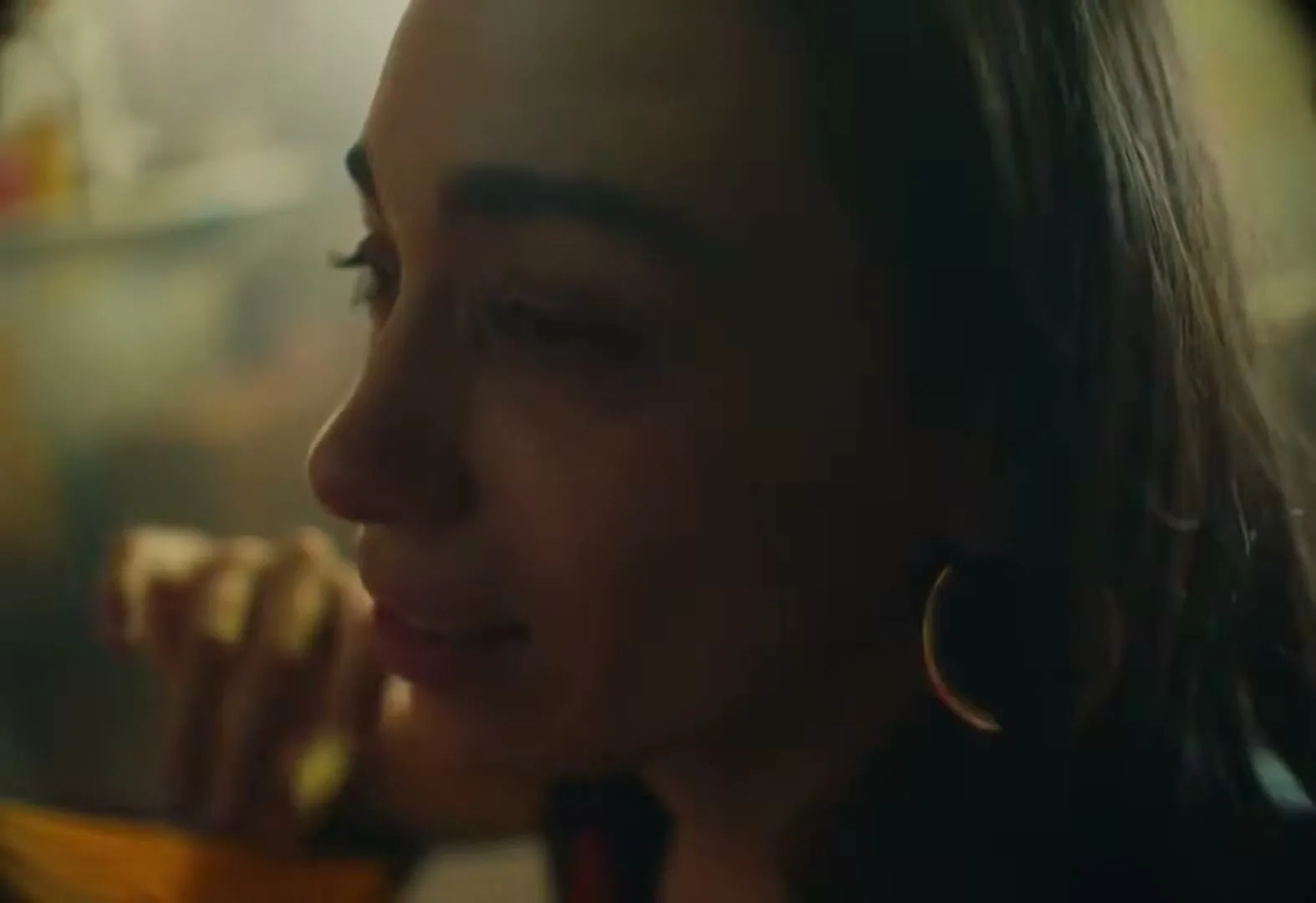 A young woman with dark hair and hoop earrings eats a taco with her hands, then smiles, in a dimly lit restaurant.