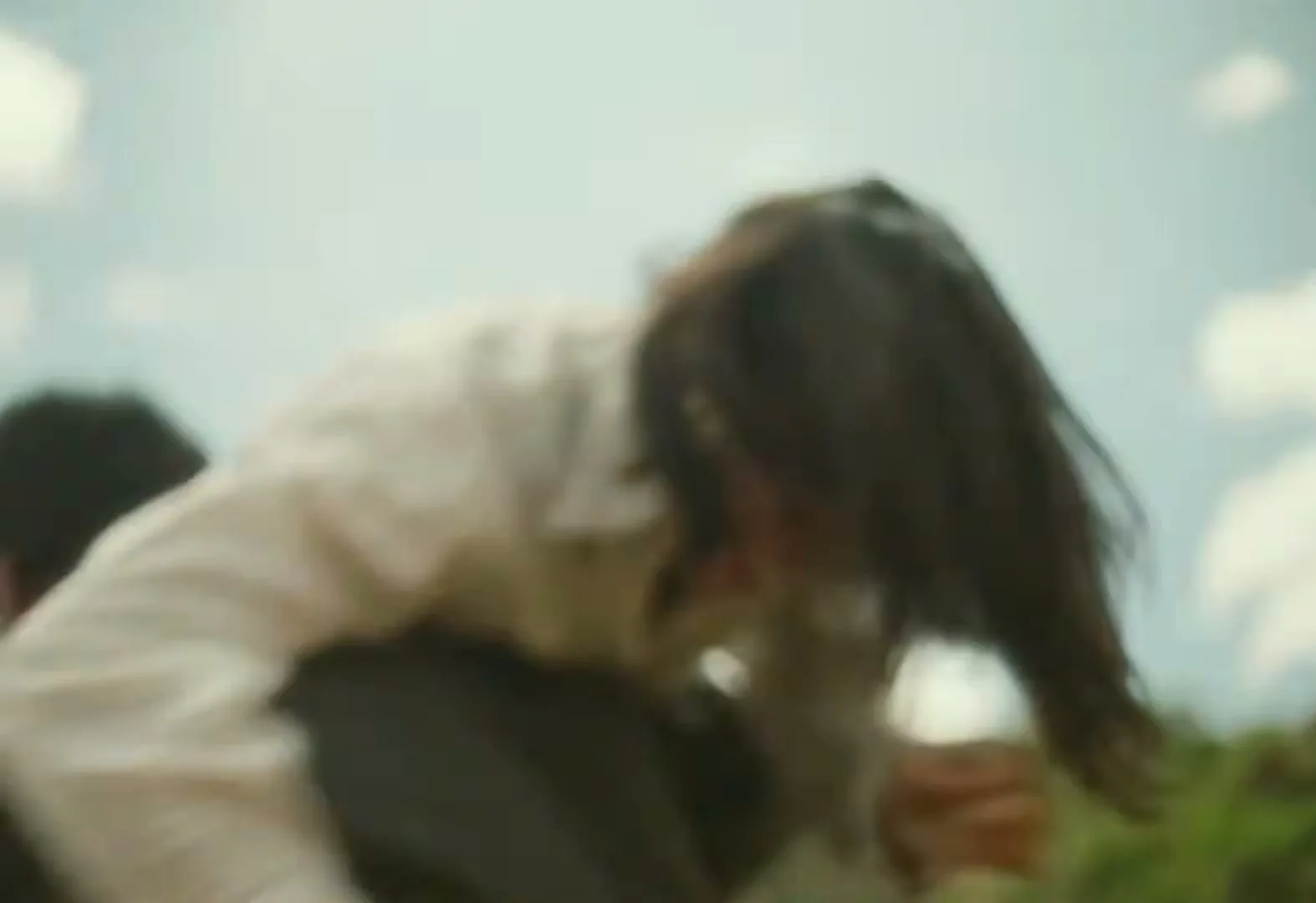 A man in a grey jacket and a woman in a white shirt struggle and fall to the ground on a grassy hillside under a cloudy sky.
