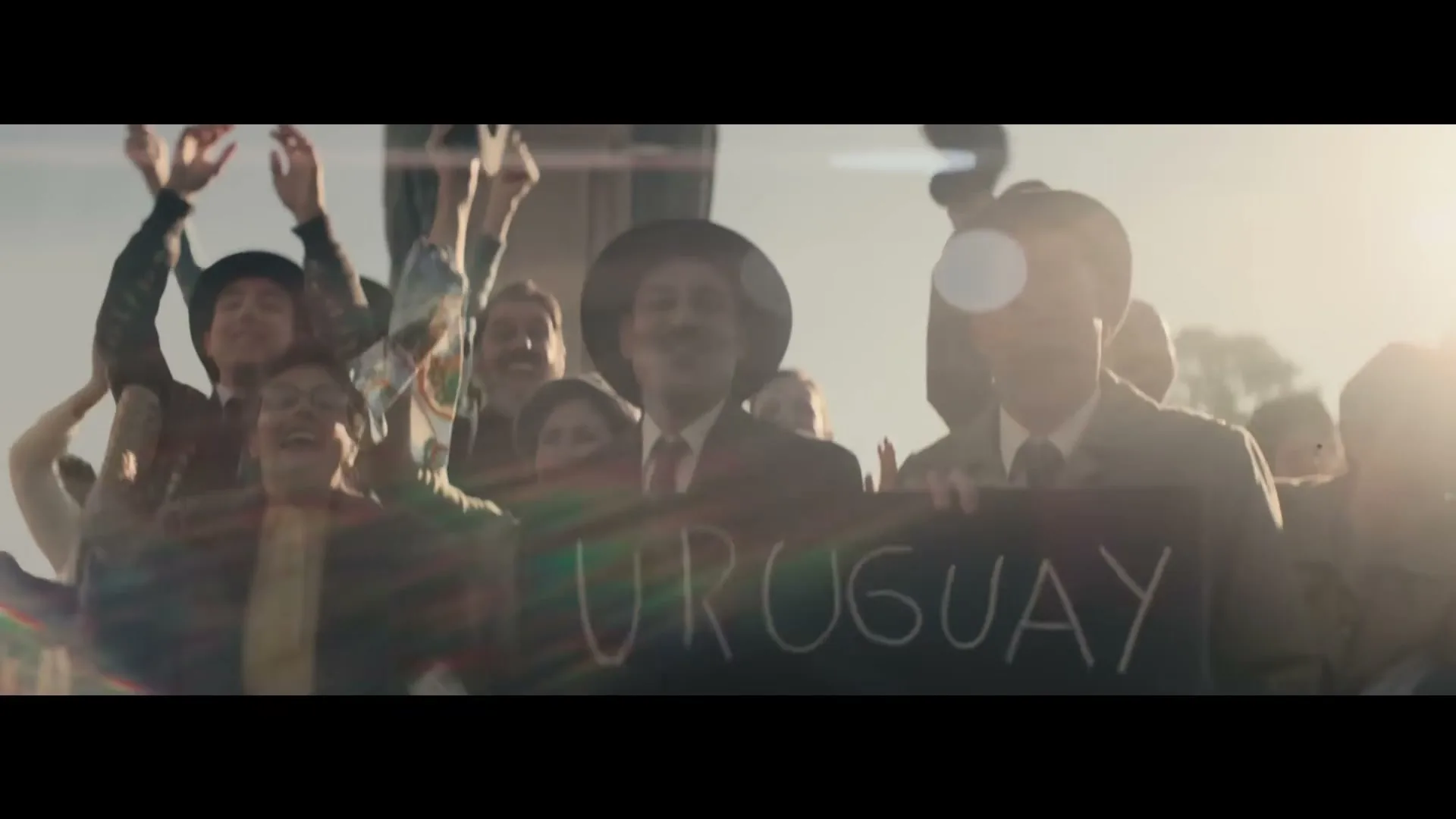 A crowd of people, including men in suits and hats, cheer and hold up a "URUGUAY CAMPEON" sign, backlit by a bright sun.