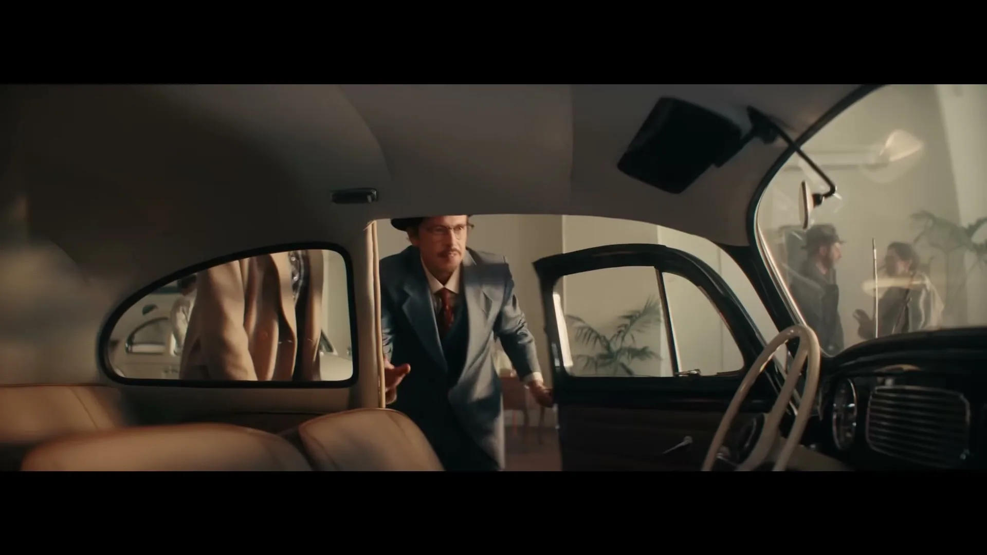 A man in a suit and hat opens the door of a vintage car, looking inside with a surprised expression in a showroom.