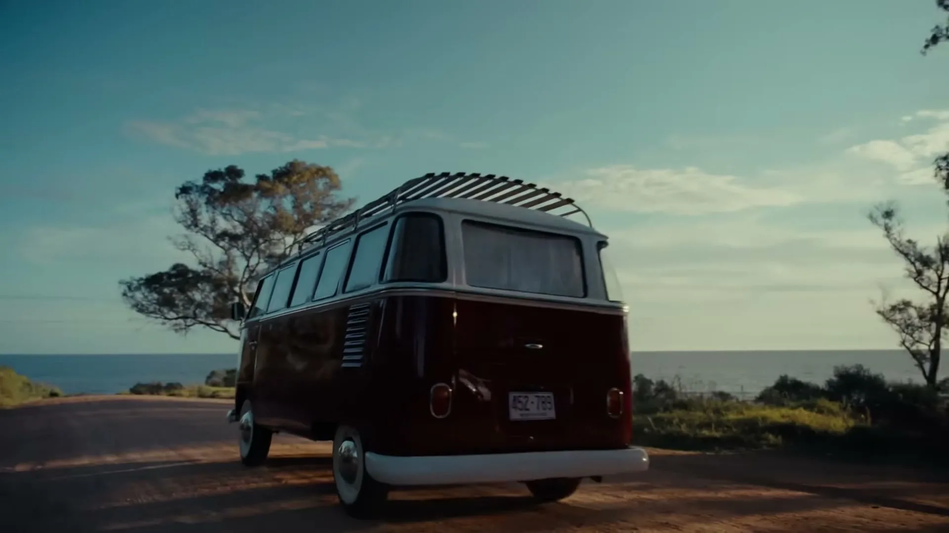A vintage red Volkswagen van drives away on a dirt road, with the ocean and blue sky visible in the background.