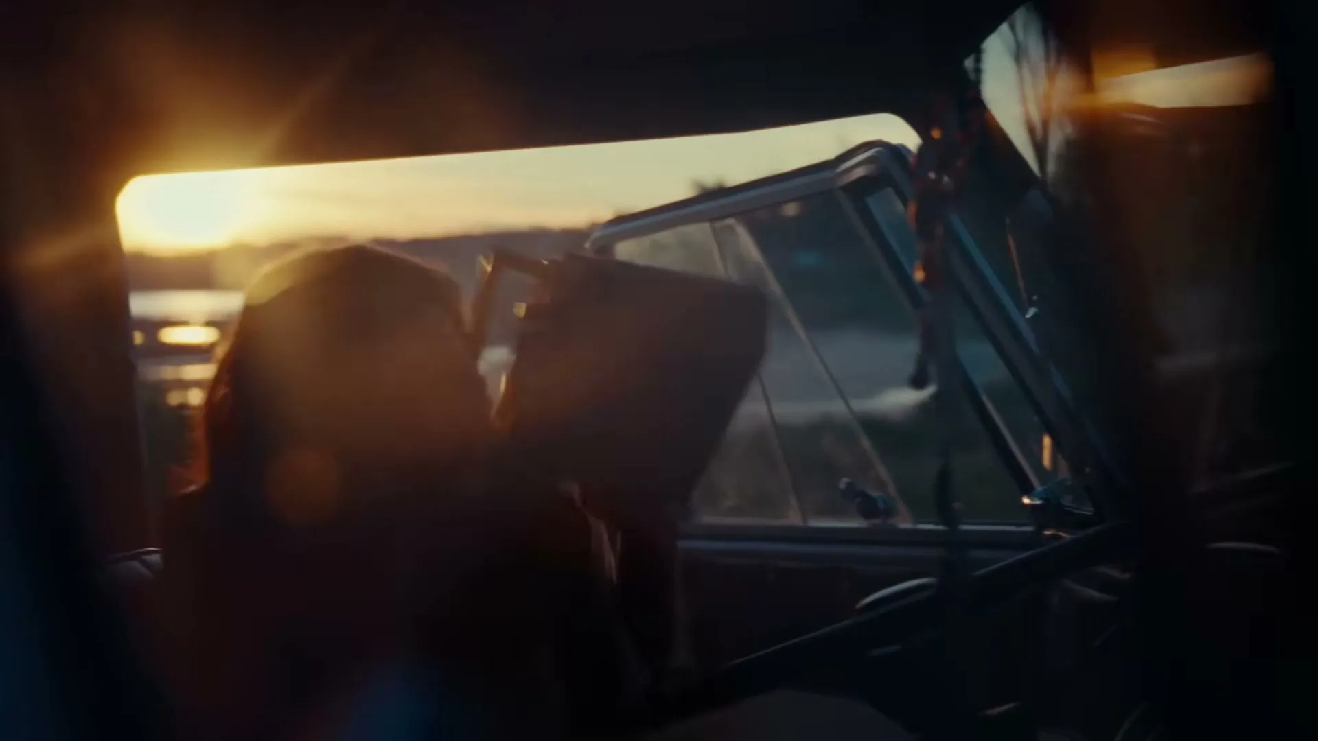 A young woman with long hair, seen from behind, is driving a vintage car at sunset, with lens flare from the sun.