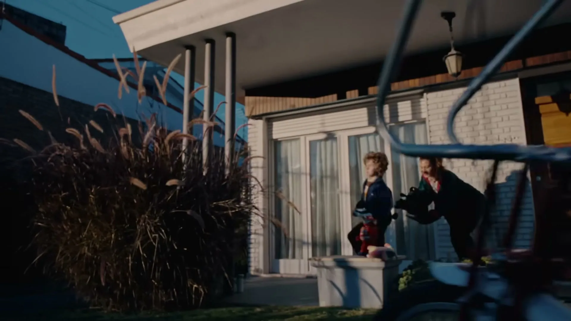 A young boy and a woman run out of a modern house, playing with water guns, as a bicycle passes in the foreground.