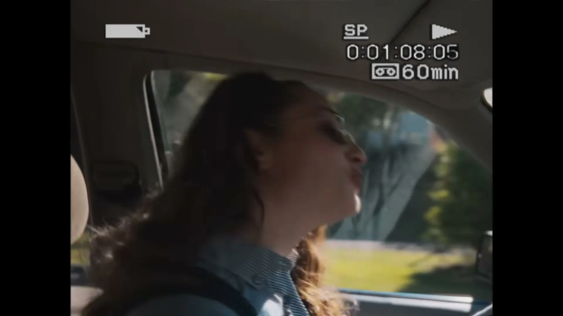 A young woman with long, curly hair and sunglasses drives a car, looking up and singing joyfully on a sunny day.