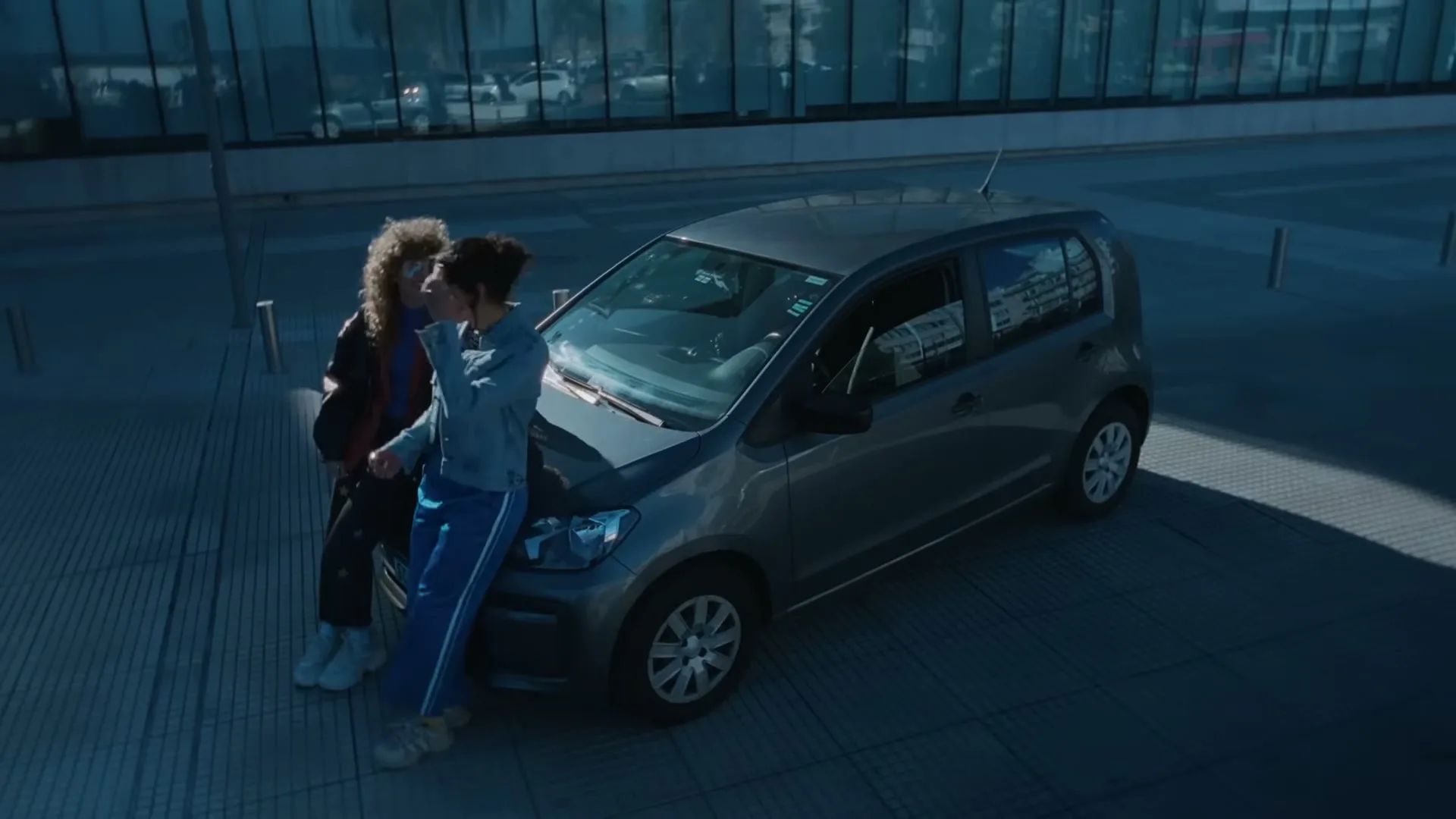 Two young women, one with curly hair and sunglasses, the other with dark hair, stand next to a gray car in a modern urban setting.