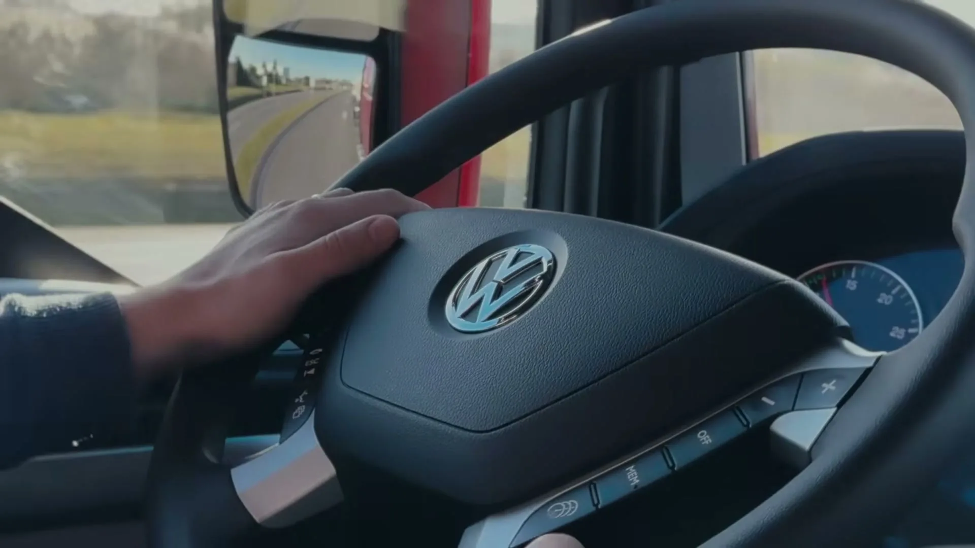 A man's hand is seen on the steering wheel of a Volkswagen truck, driving on a highway during the day.