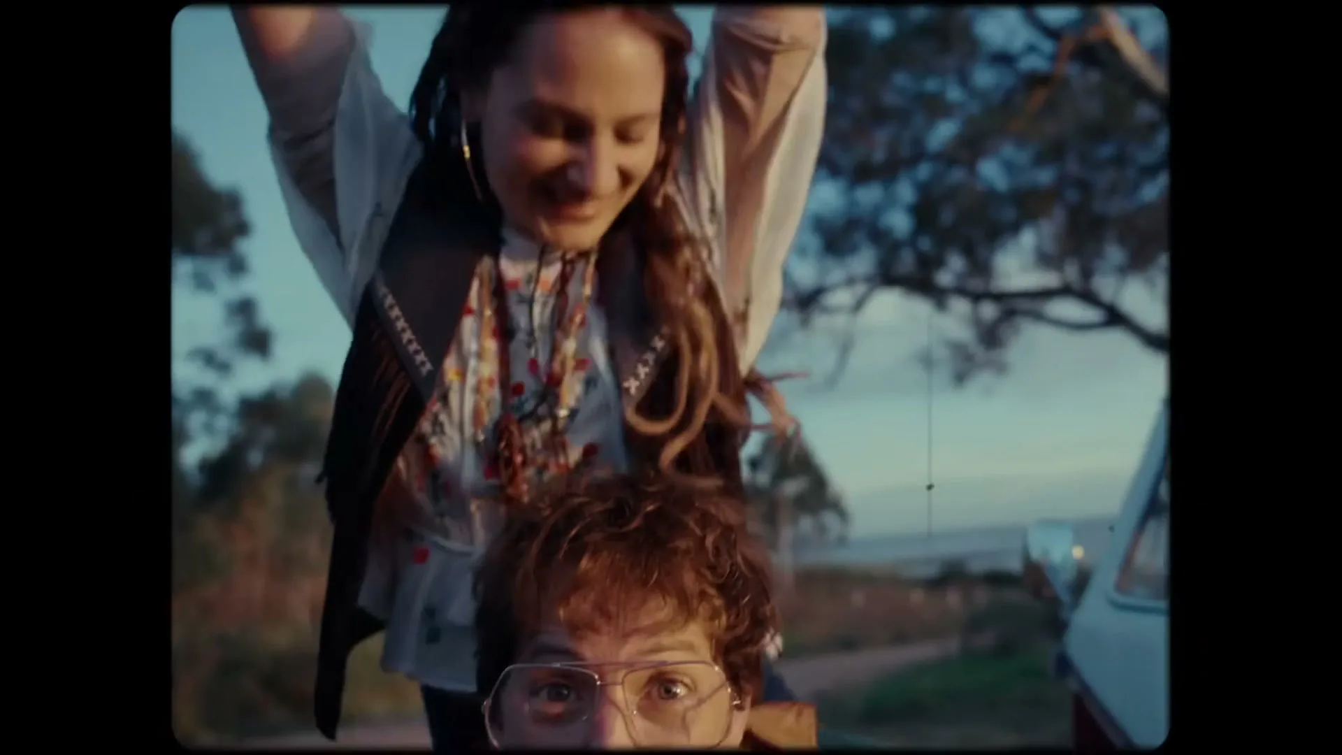 A young man with curly hair and glasses looks up from a car, while a woman with dreadlocks and a vest stands behind him, raising her arms in joy.