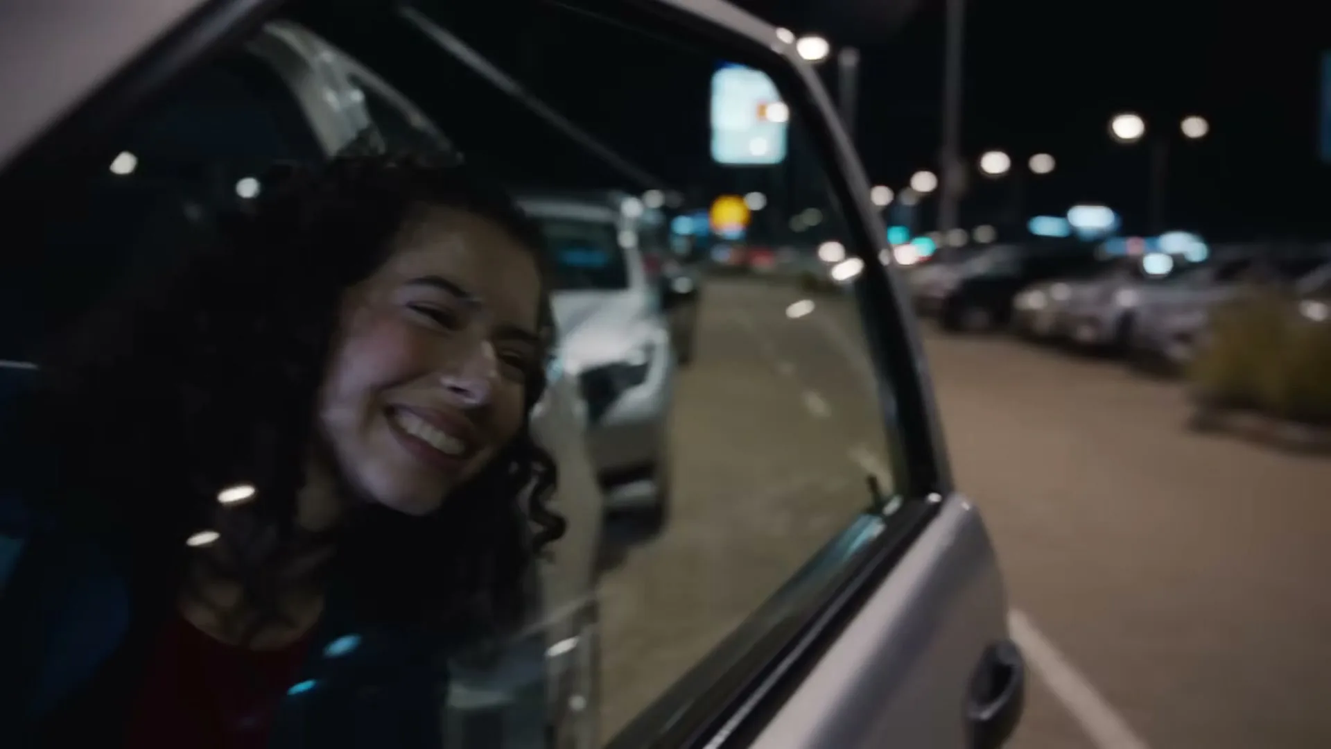 A young woman with curly hair smiles happily, looking out the open car window at night in a parking lot.