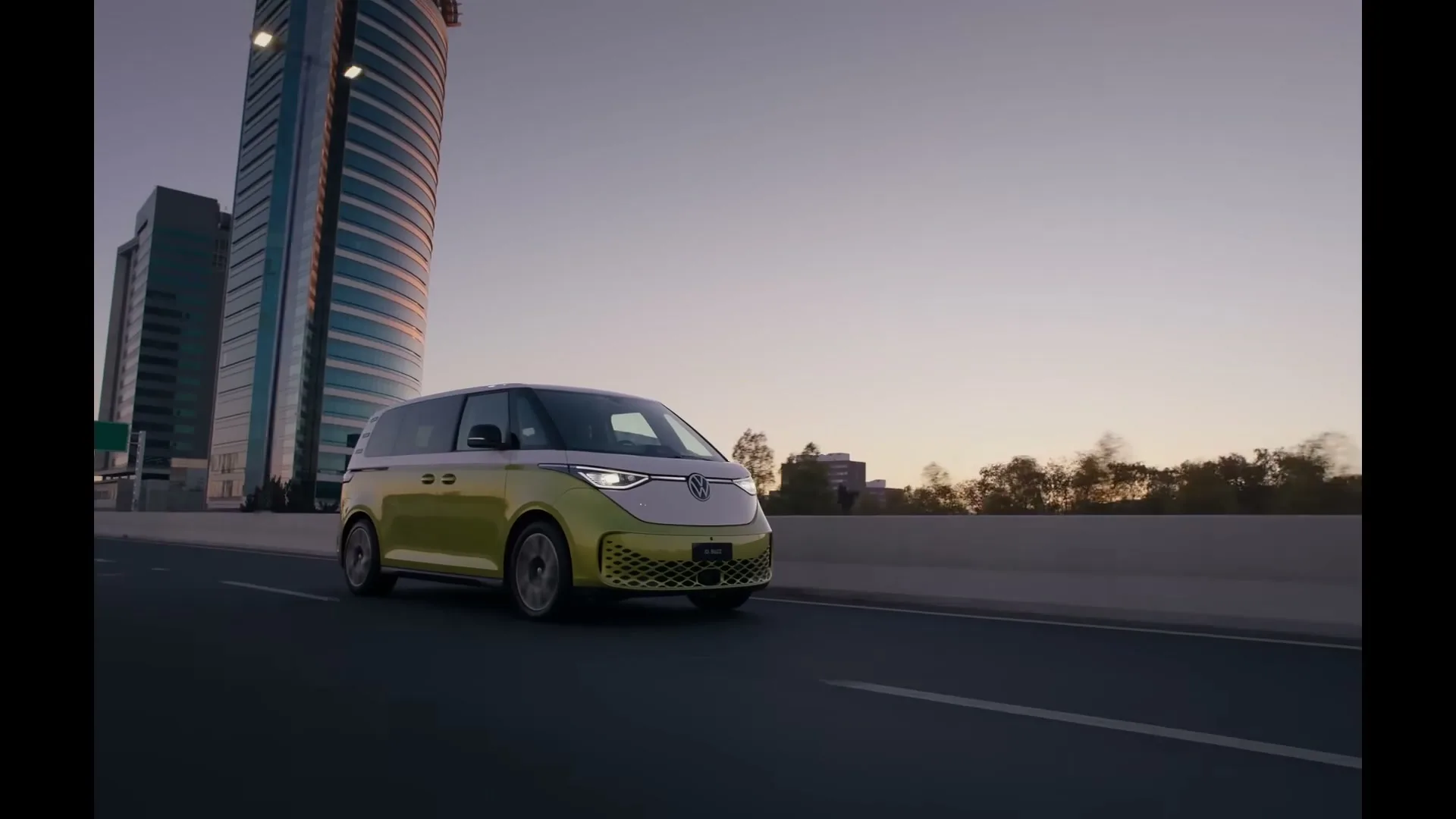 A yellow and white Volkswagen ID. Buzz electric van drives on a highway next to a modern skyscraper at dusk.