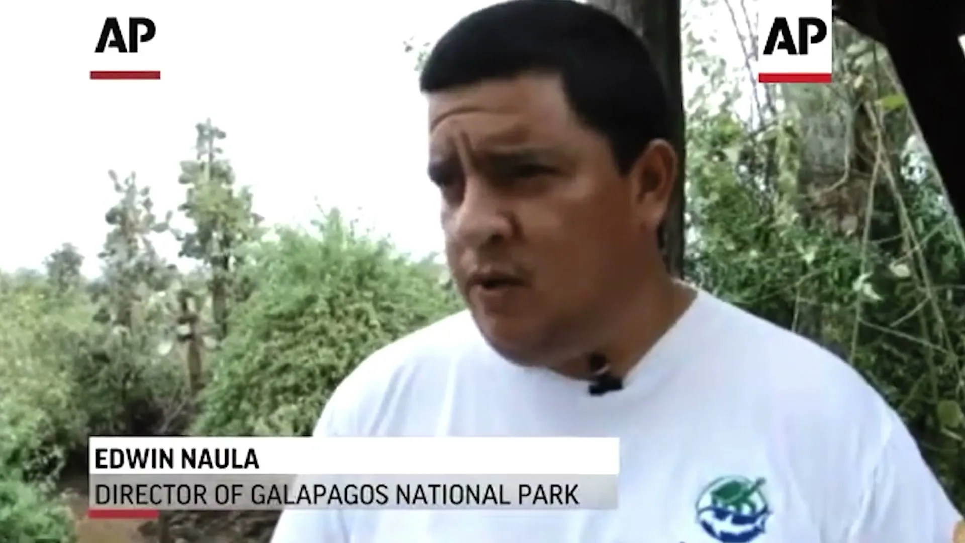 A man in his 40s, wearing a white t-shirt, speaks to the camera in an outdoor setting with trees in the background.