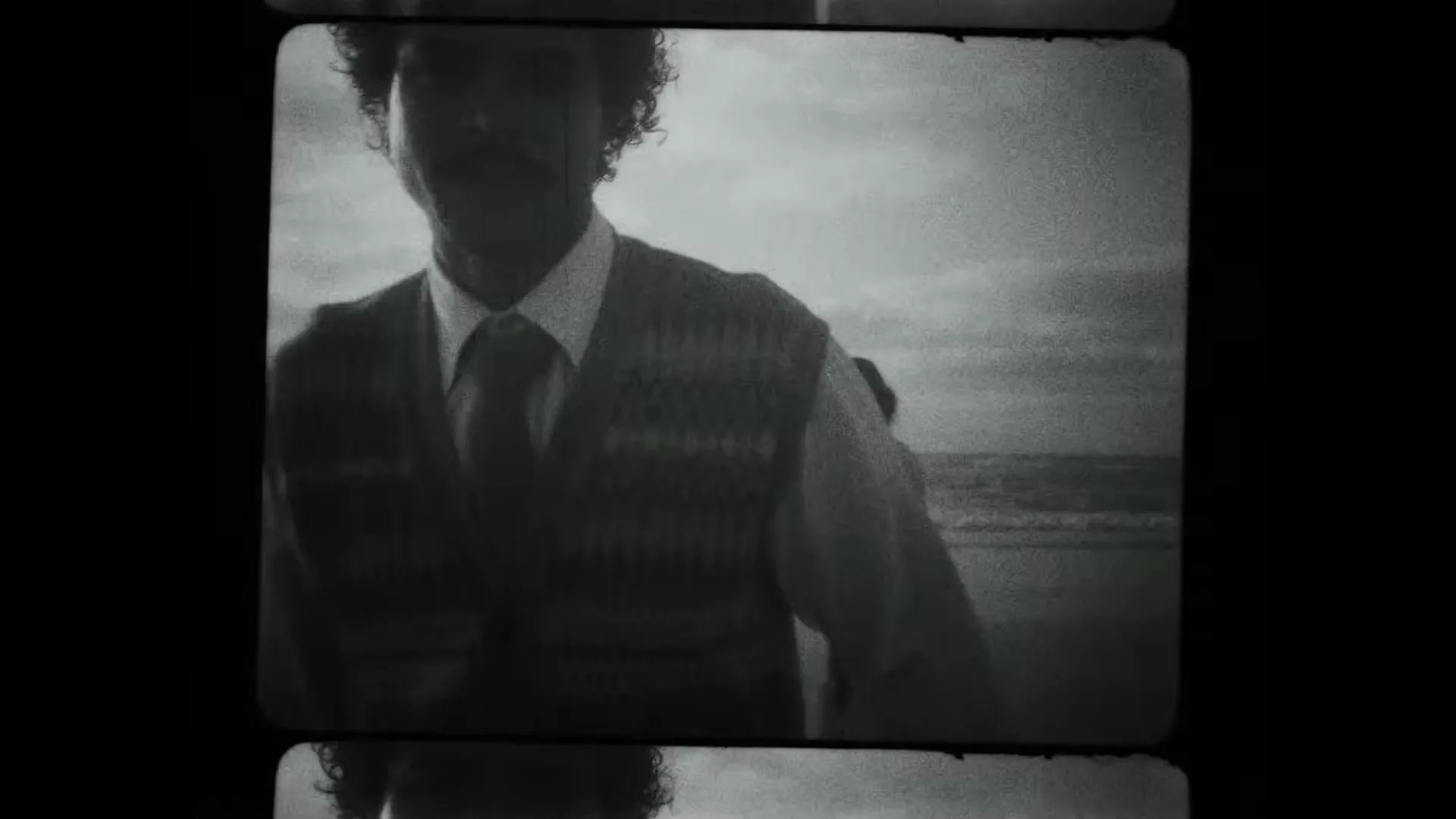 A man with curly hair and a mustache, wearing a sweater vest and tie, walks on a beach with other people in a grainy, black and white film.