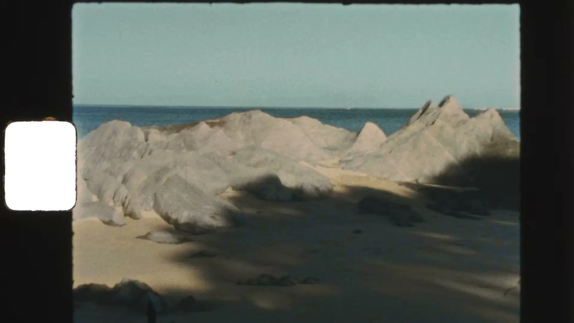 A wide shot shows a sandy beach with large, light-colored rock formations under a clear blue sky, with the ocean visible in the distance.
