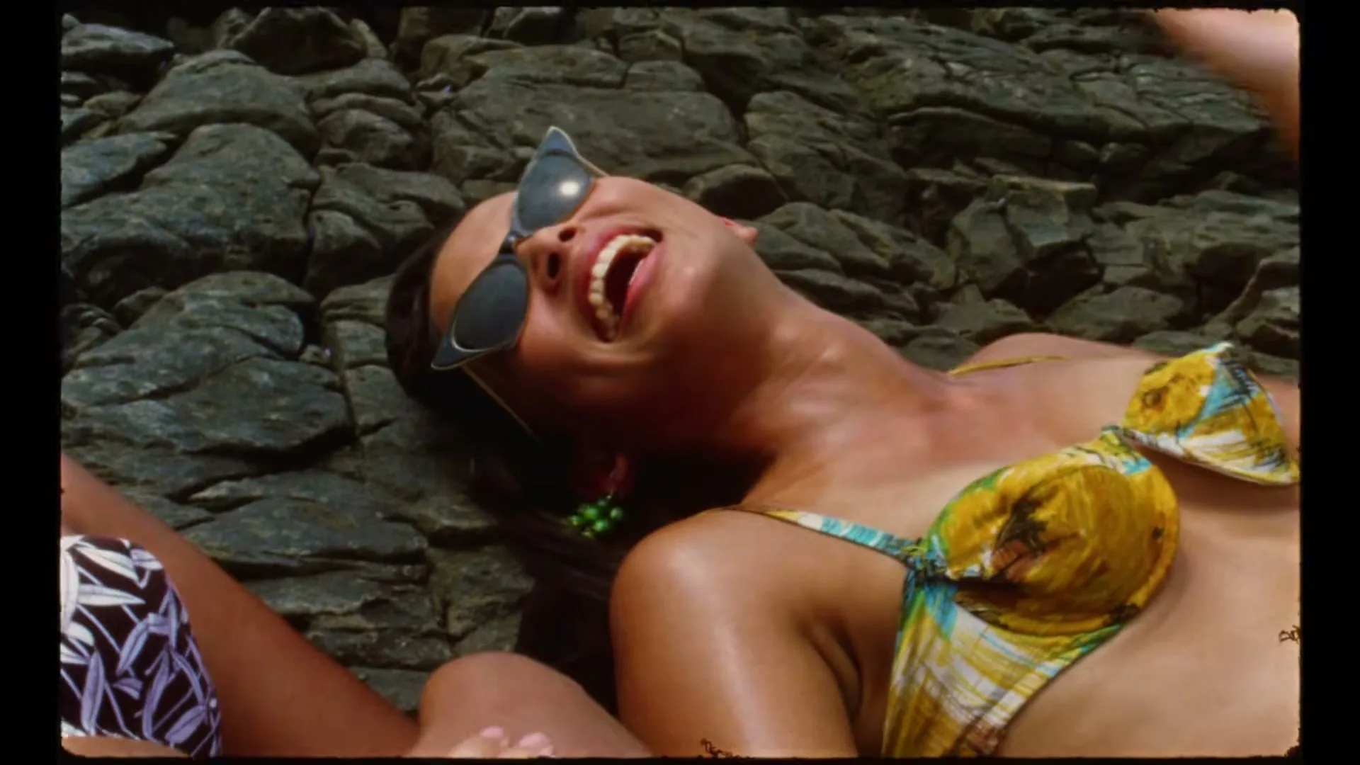 A woman in sunglasses and a bikini laughs joyfully while lying on dark rocks, with another person's arm visible beside her.
