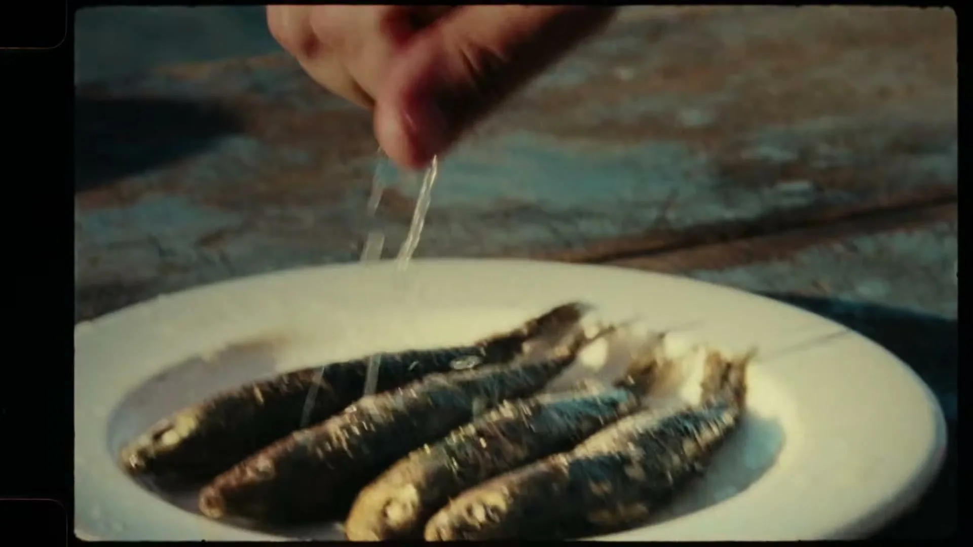 A person's hand sprinkles salt over four small, cooked fish arranged on a white plate, on a rustic wooden surface.