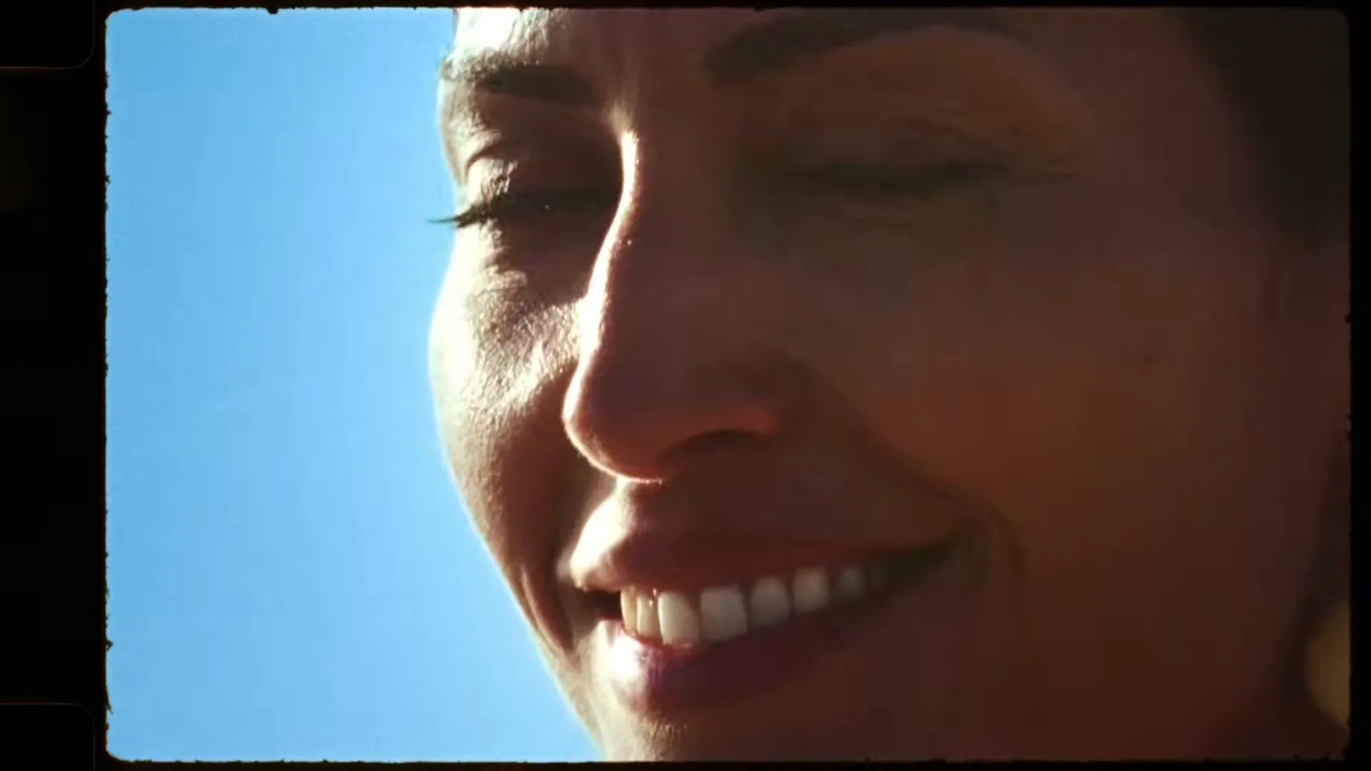 A woman with dark hair and tanned skin smiles, looking up and to the right, against a bright blue sky.