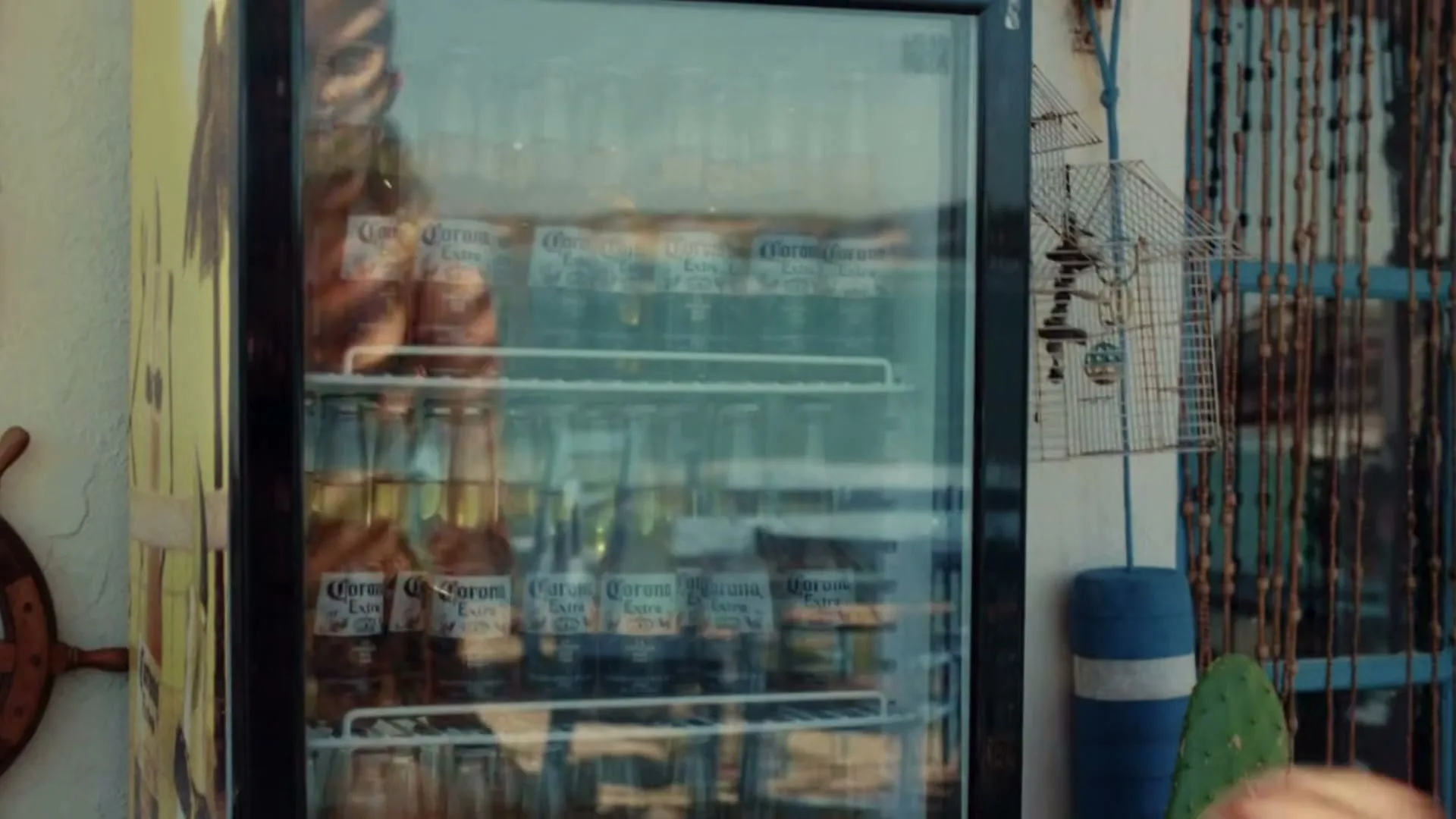 A woman in a swimsuit reaches for a Corona beer from a glass-fronted refrigerator on a sunny patio.