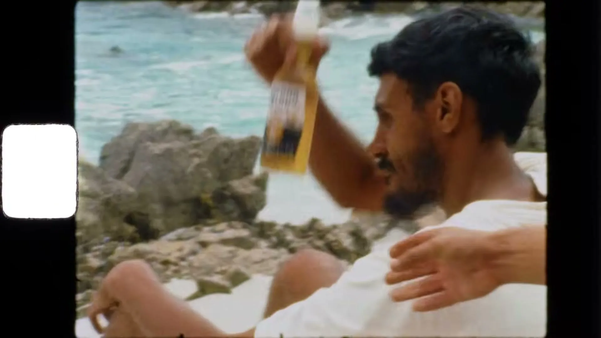 A man with dark hair and a beard, wearing a white shirt, holds a Corona beer bottle on a rocky beach with turquoise water.