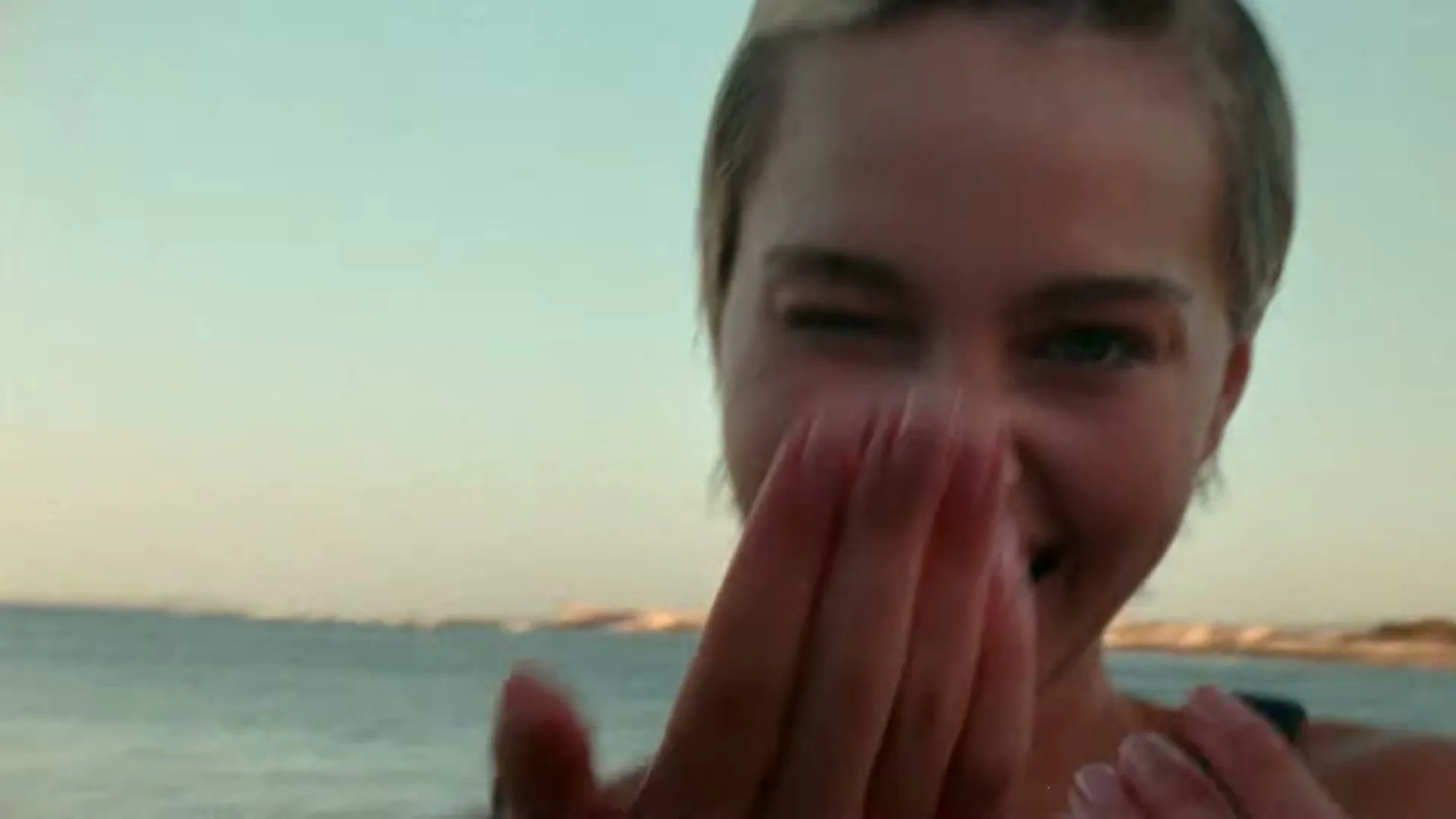 A young woman with short blonde hair winks and smiles at the camera, beckoning with her hand on a beach at golden hour.