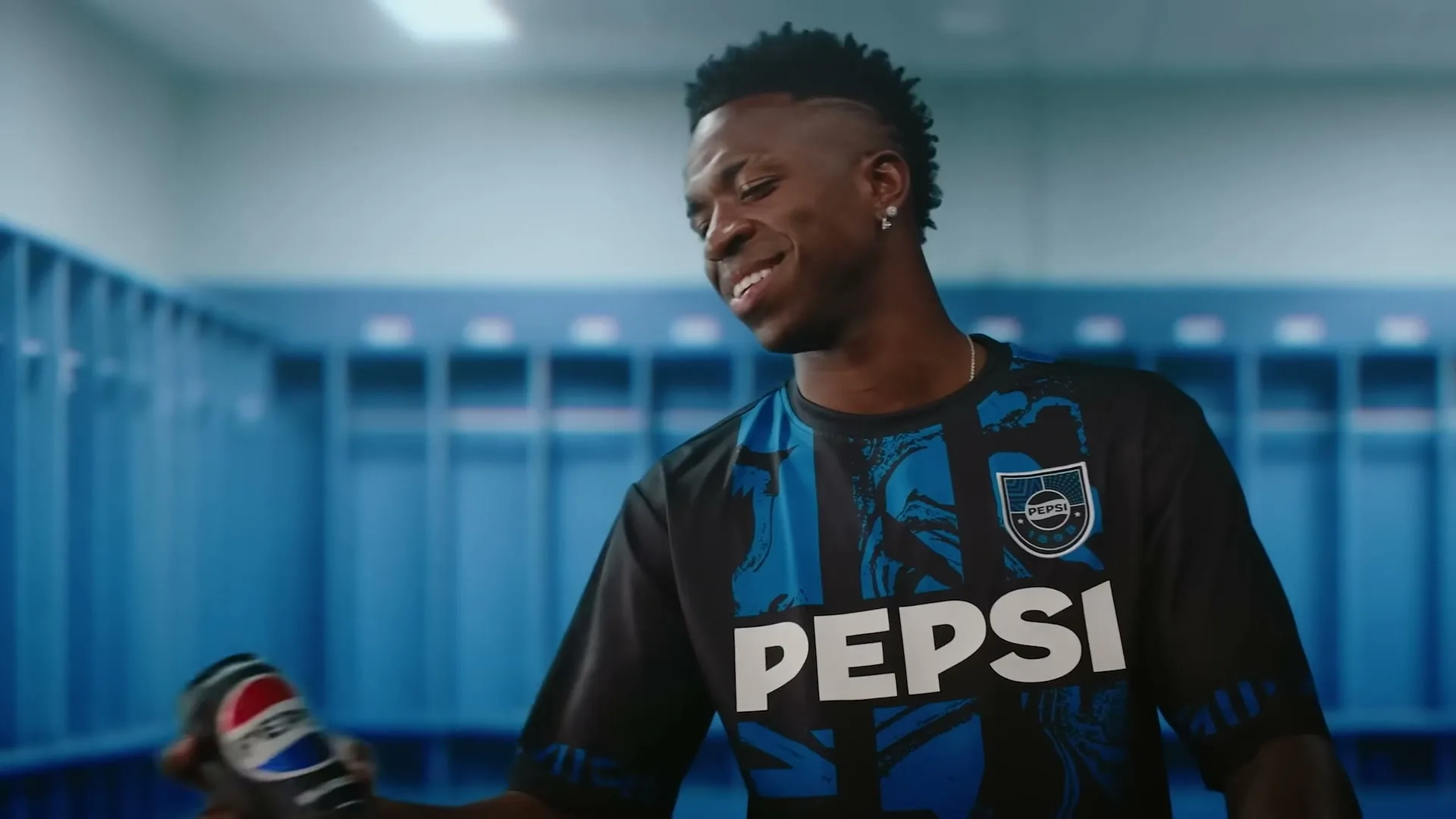 A male soccer player in a black and blue jersey smiles while holding a Pepsi can in a blue locker room.