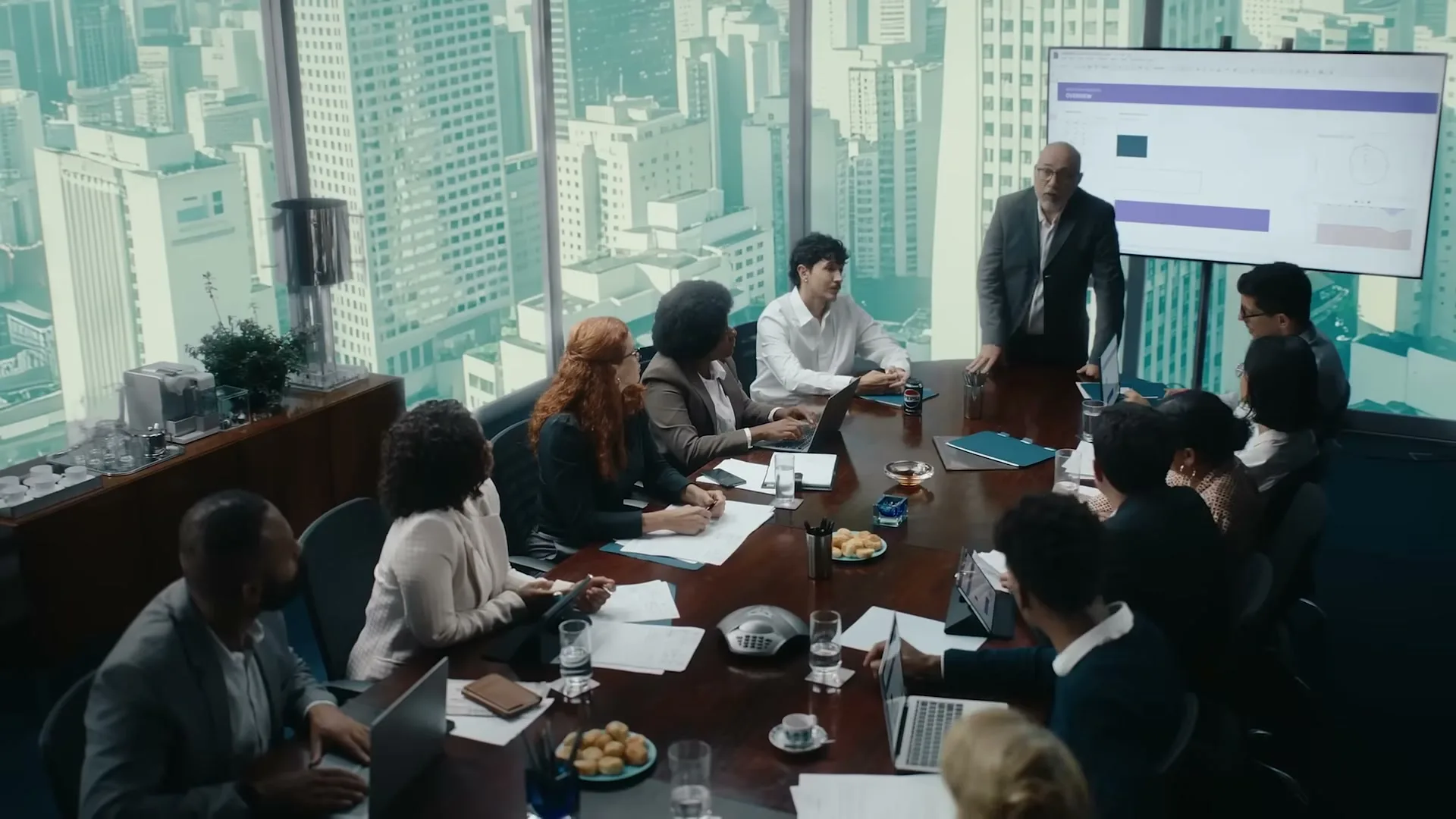 A diverse group of business people are seated around a large conference table in a high-rise office, listening to a male presenter.