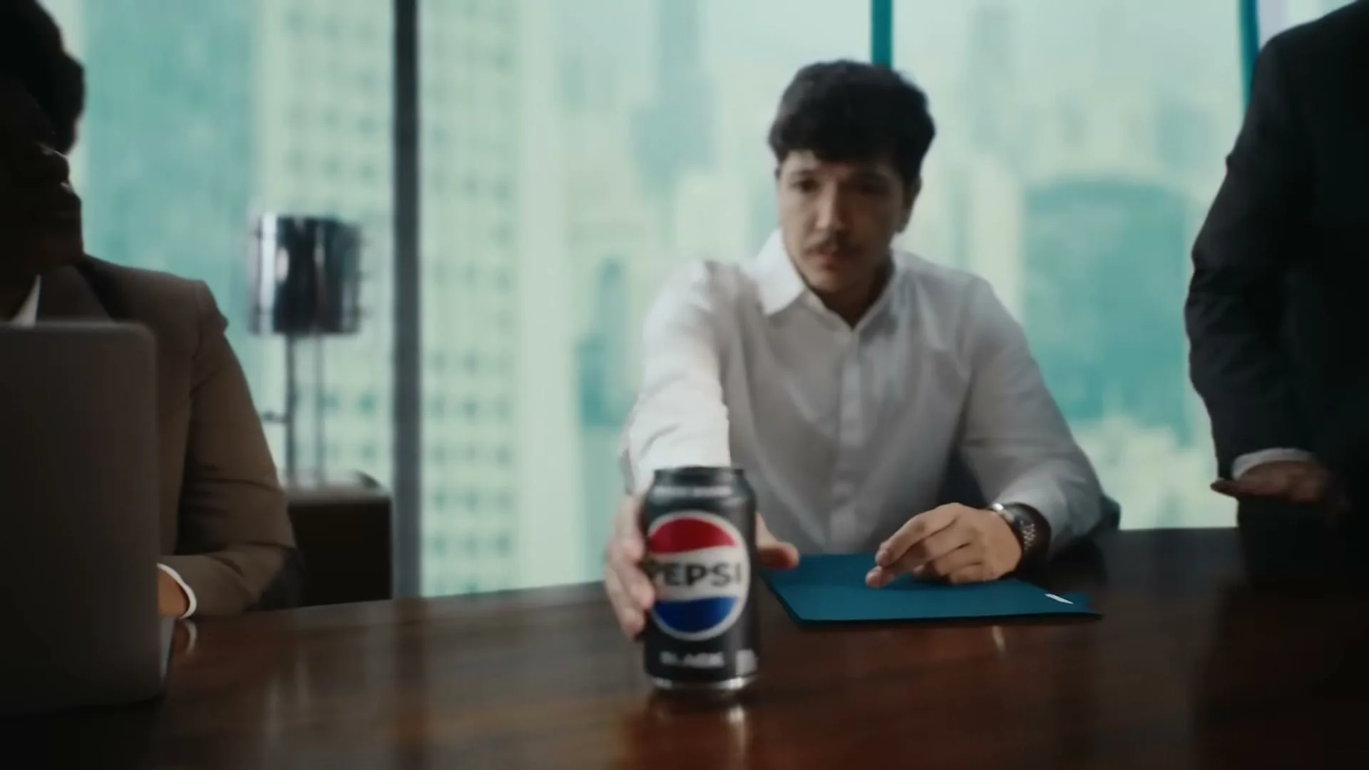 A man in a white shirt reaches for a can of Pepsi Black on a conference table, with blurred colleagues and city buildings in the background.