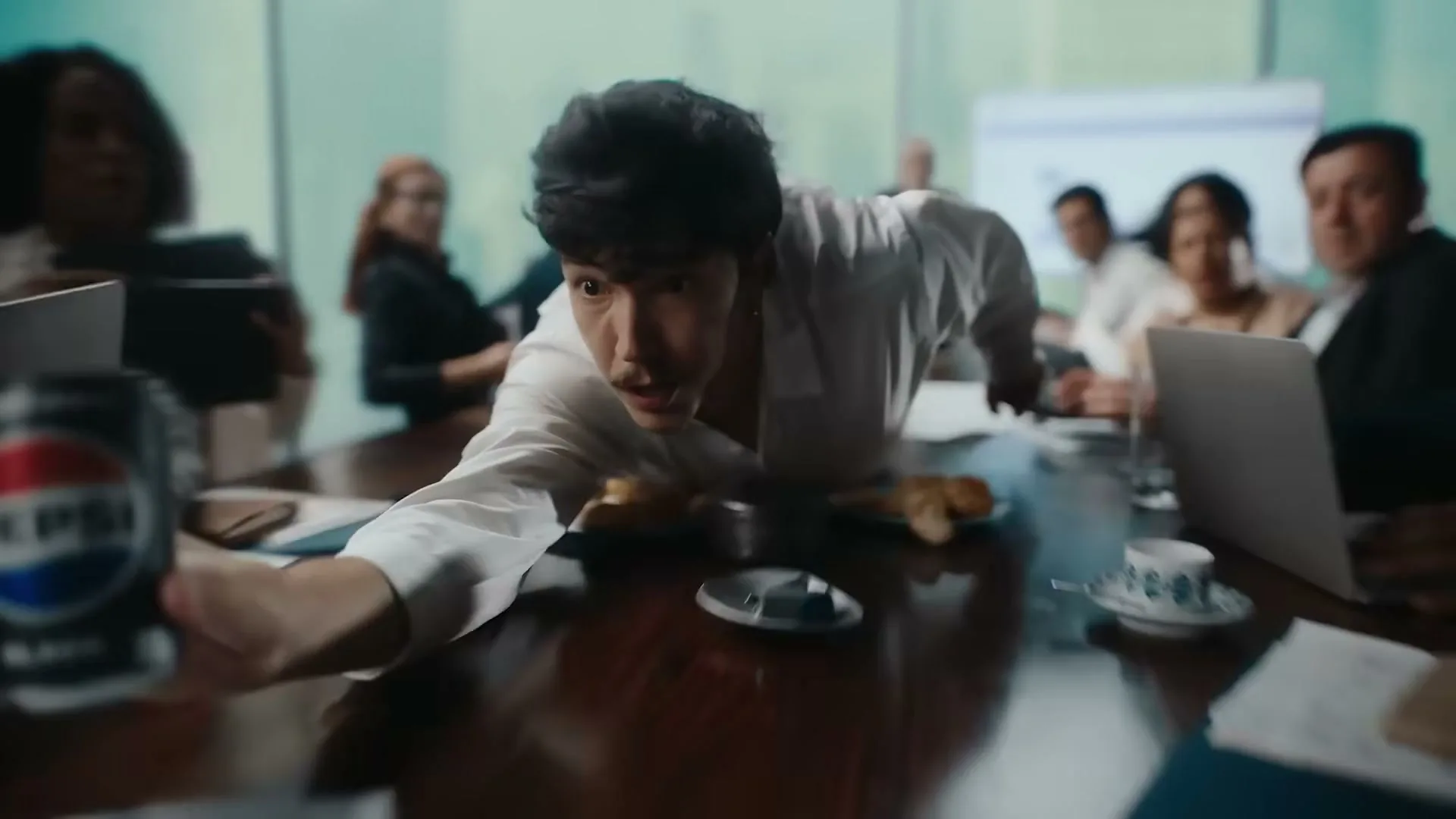 A young man in a white shirt reaches for a Pepsi Black can on a conference table, surrounded by colleagues in a meeting room.