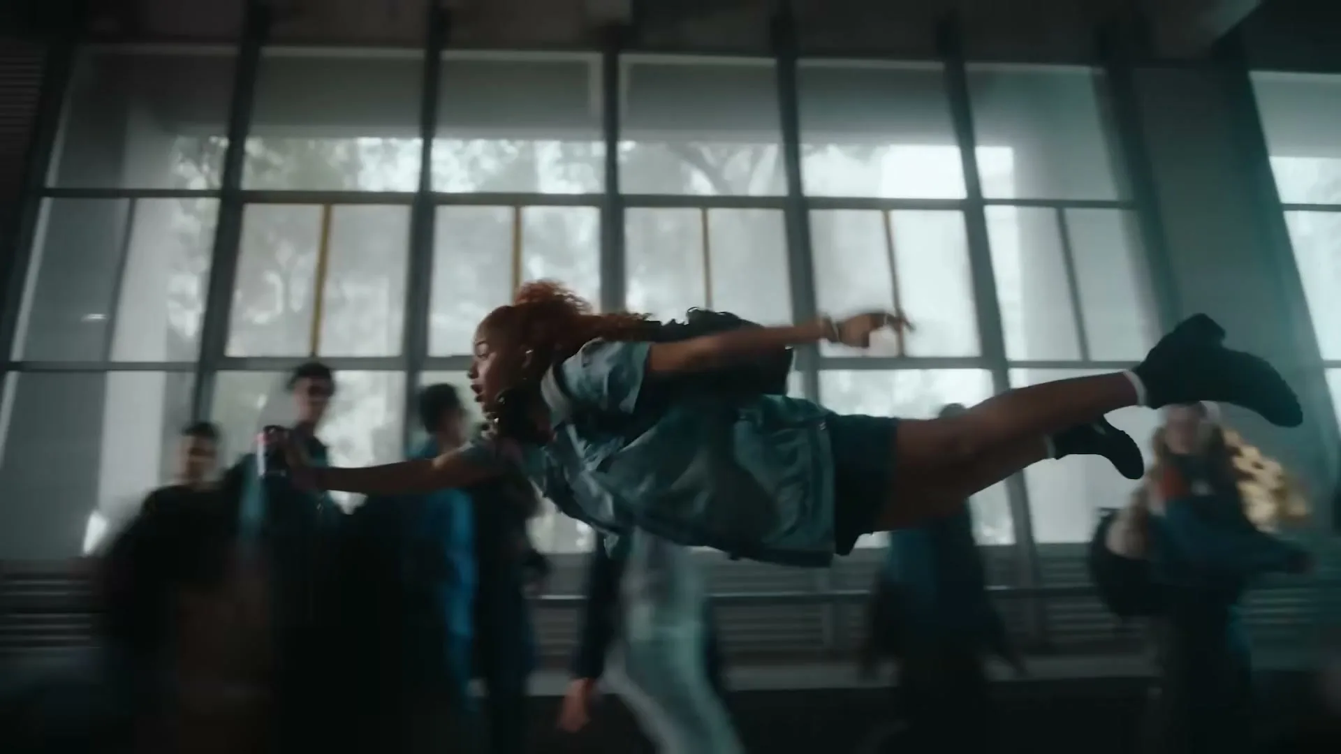 A young woman with curly red hair, wearing a denim jacket and shorts, floats horizontally through a crowded modern building with large windows, holding a phone.