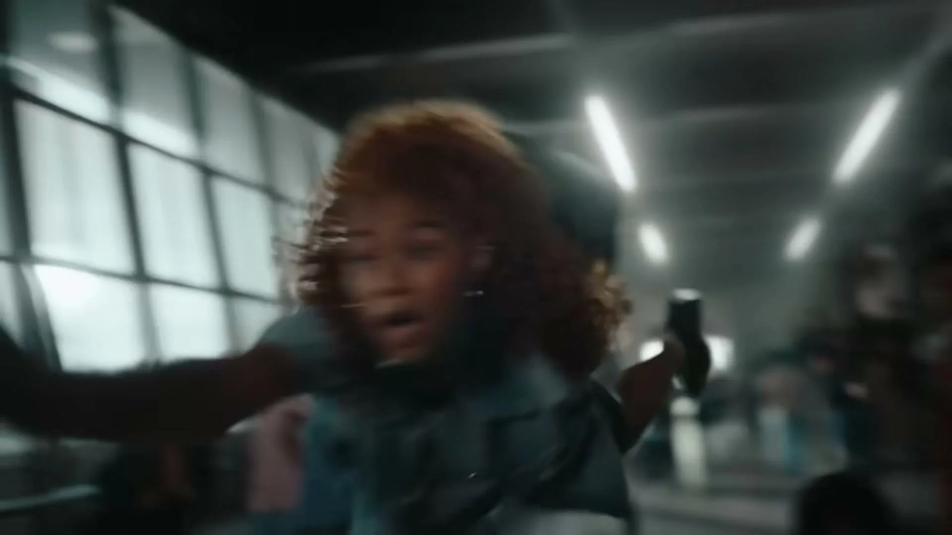 A young woman with curly hair in a denim jacket is falling backwards through a crowded, modern building interior with large windows and bright lights.