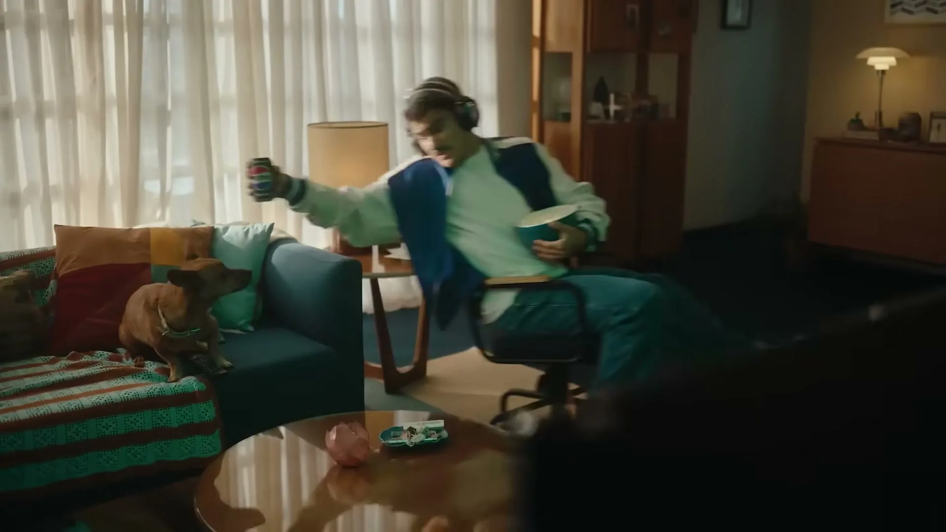A young man in a blue and white jacket, wearing headphones, is playing a video game in a living room, holding a Pepsi can and a bowl of popcorn, while a small dog sits on the couch.