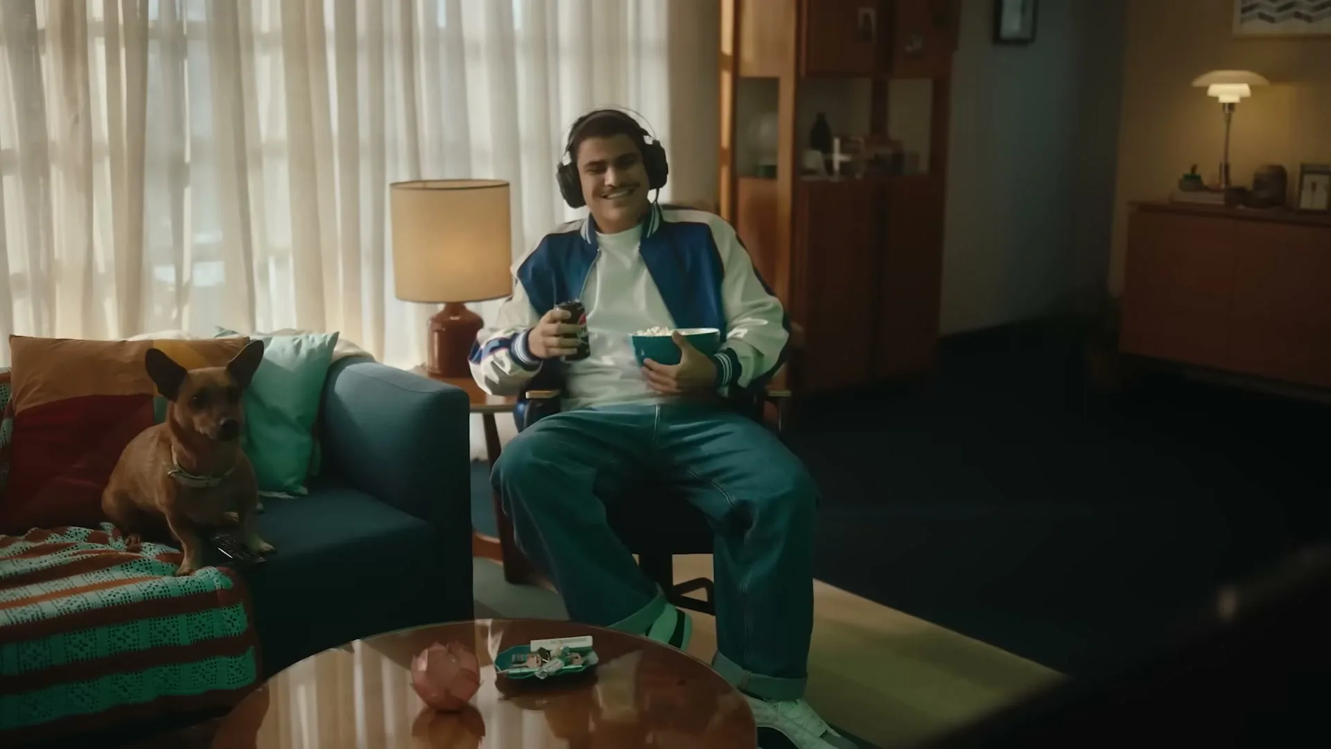 A young man in a varsity jacket and headphones sits in a living room, holding popcorn and a Pepsi can, smiling at the camera with a dog on the couch next to him.