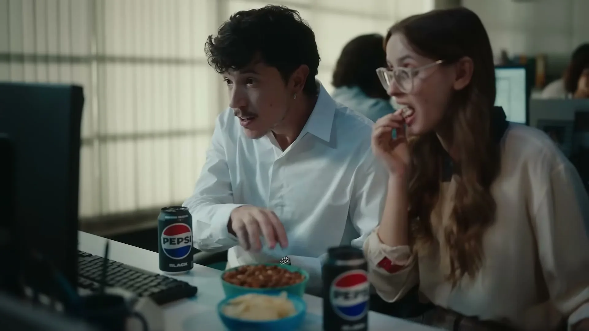 A young man and woman in an office cubicle are looking at a computer screen, eating snacks, and drinking Pepsi Black.