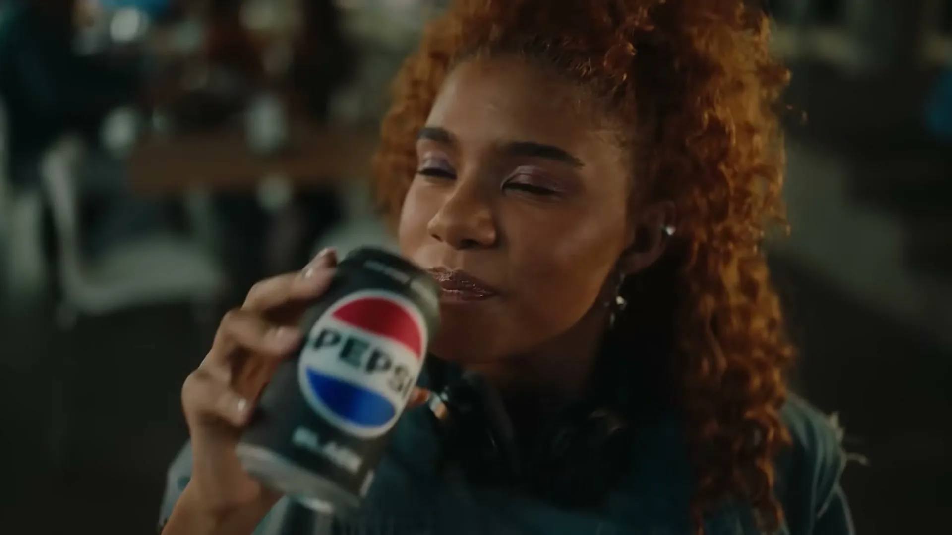 A young woman with curly red hair drinks from a can of Pepsi Black, then looks up, in a dimly lit interior.