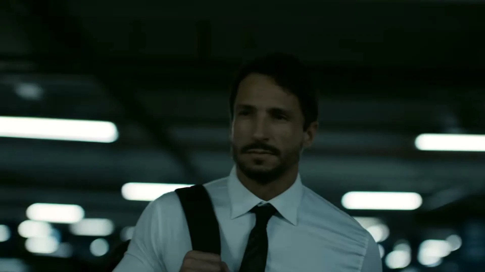 A man in his 30s, wearing a white shirt and black tie, walks through a dimly lit parking garage with a neutral expression.