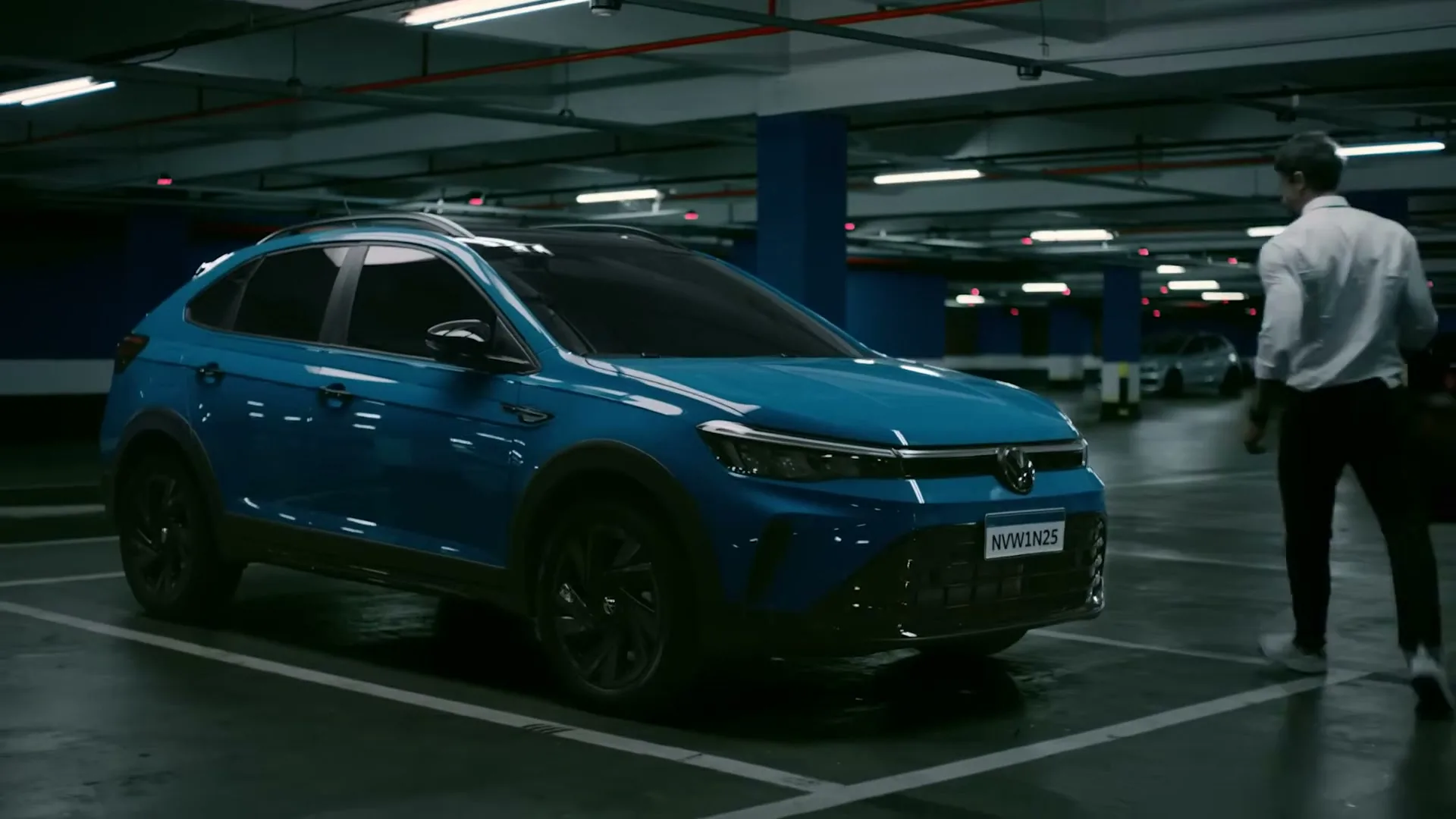 A man in a white shirt and dark pants walks towards a blue Volkswagen car parked in an underground parking garage.