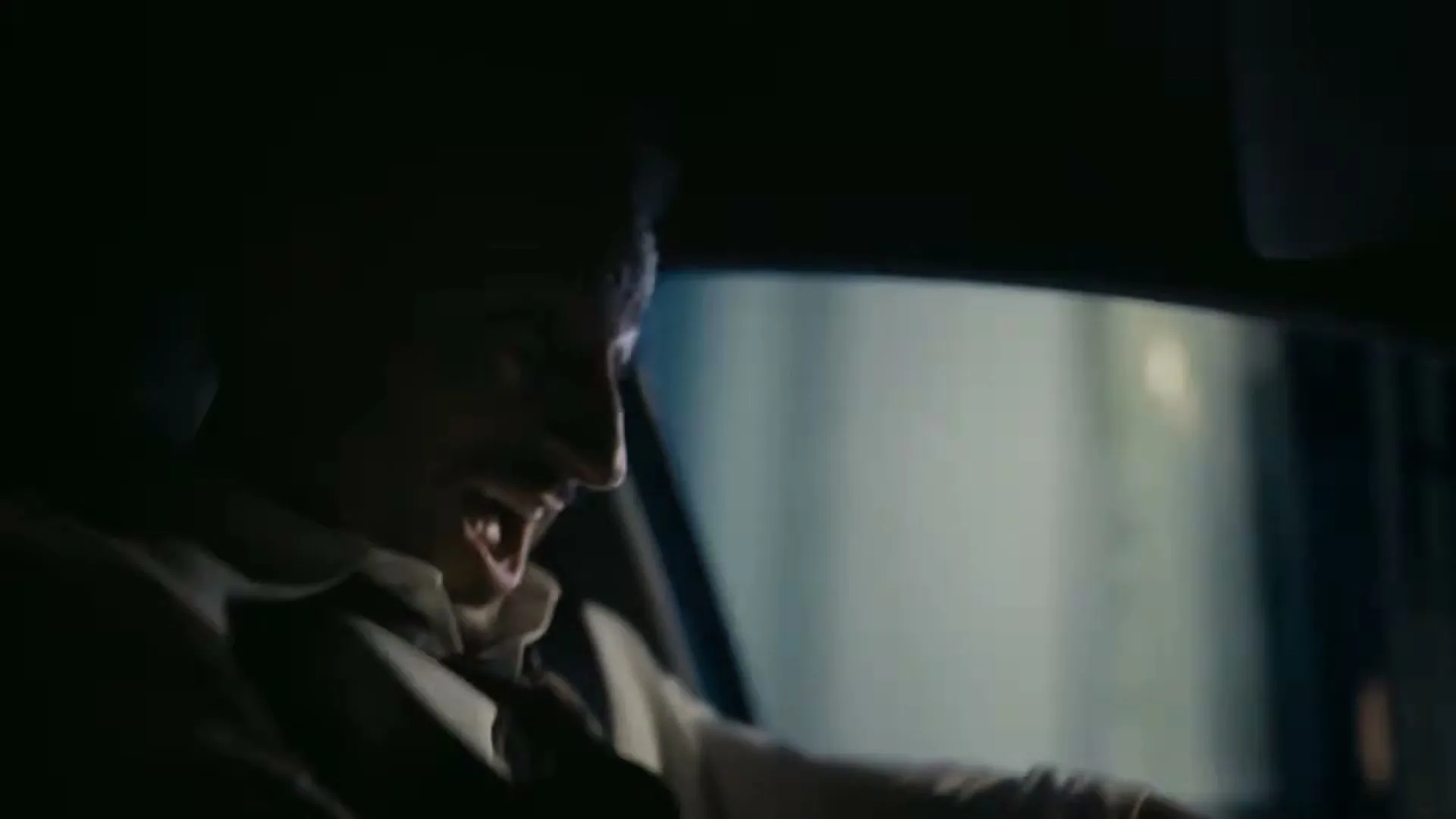 A man in his 30s, wearing a white shirt and tie, is seen from the side in a car, screaming with his mouth wide open, as if on a rollercoaster.