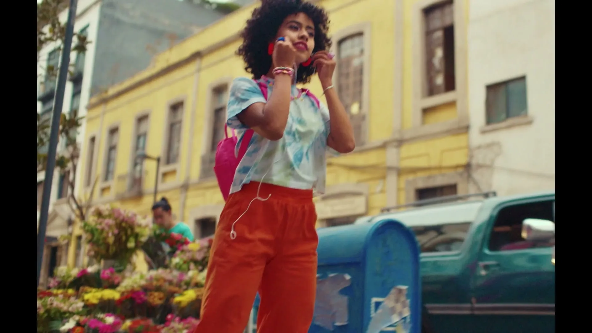 A young woman with curly hair, wearing a tie-dye shirt and orange pants, roller skates down a city street, smiling and adjusting her headphones.