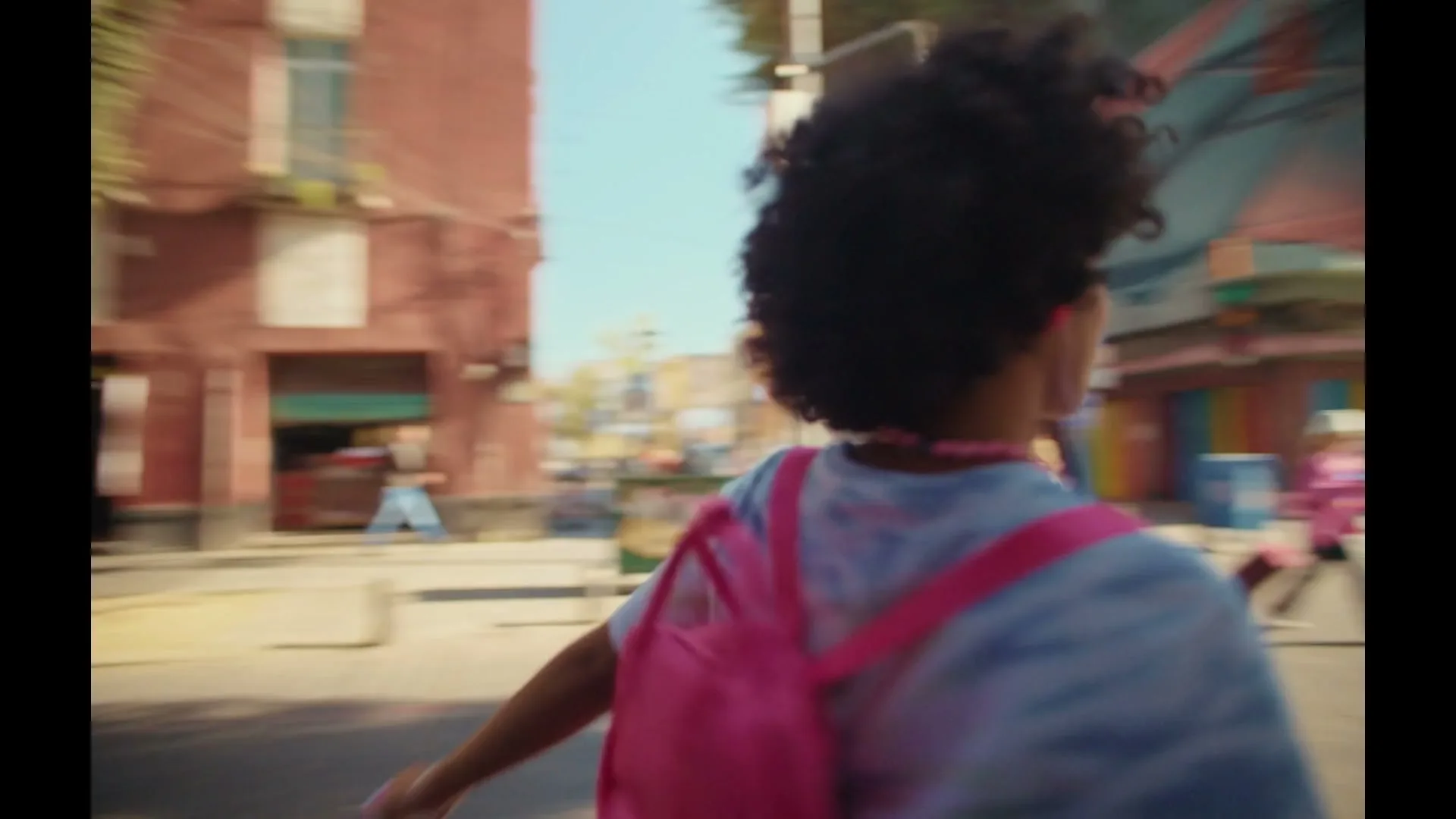 A young woman with curly hair and a pink backpack runs down a sunny city street, looking over her shoulder.