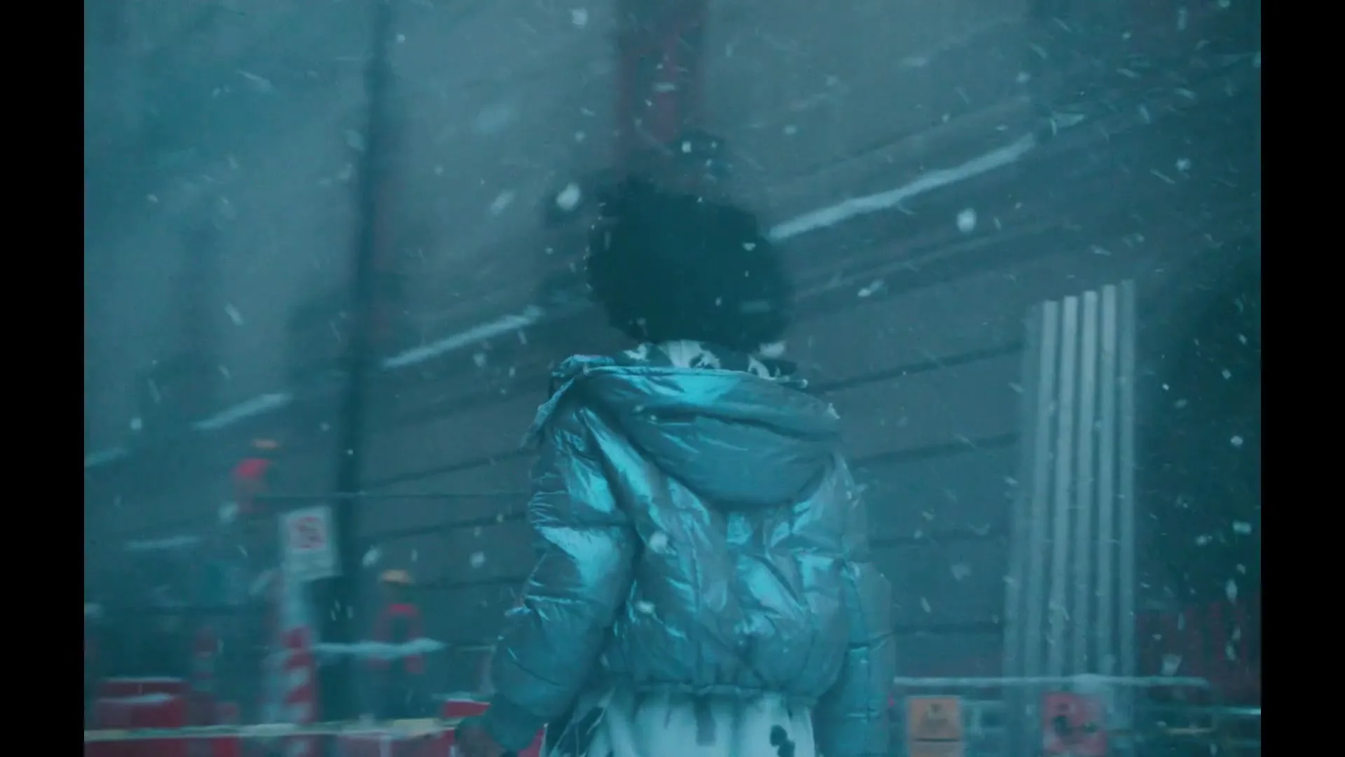 A woman with curly hair drinks from a Pepsi Black can while walking through a snowy, abandoned city street with construction barriers.