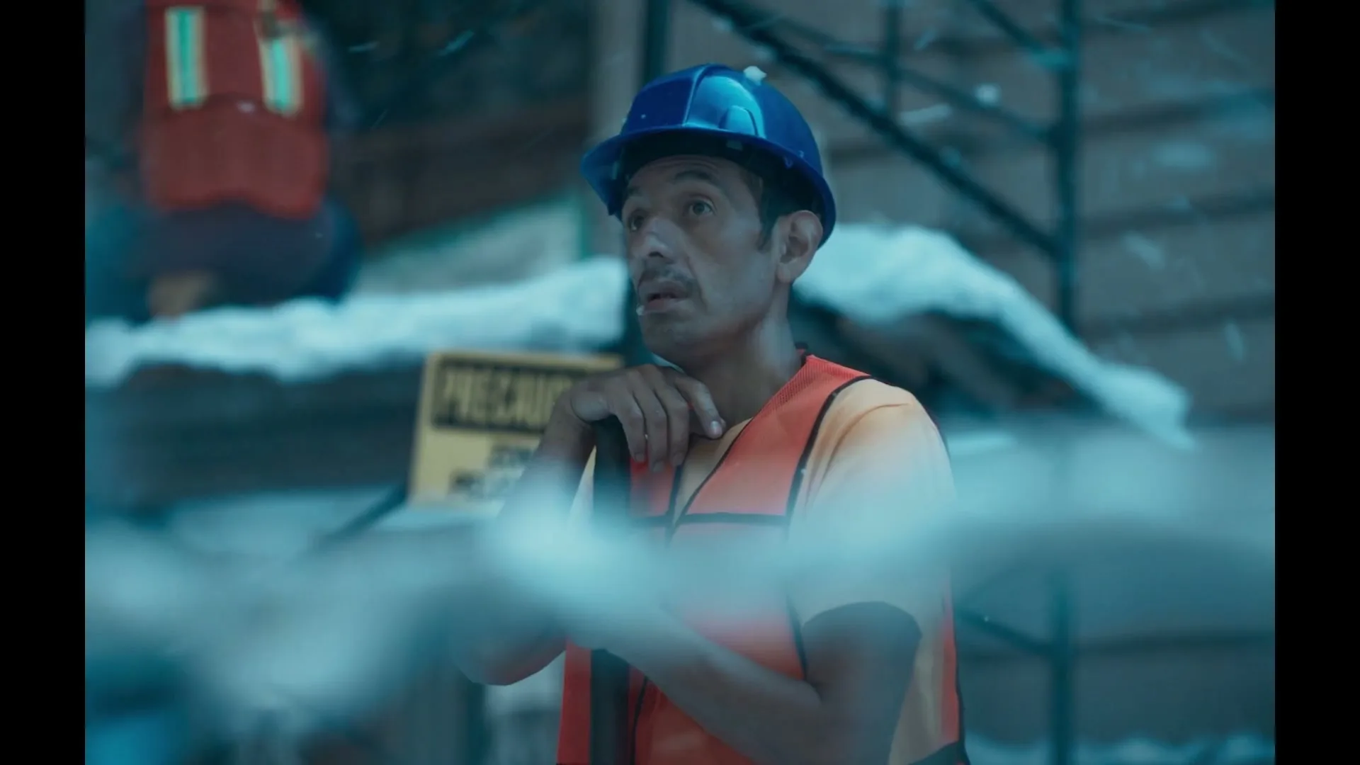 A construction worker in a blue hard hat and orange safety vest stands in a snowy, cold environment, looking around with a concerned expression.