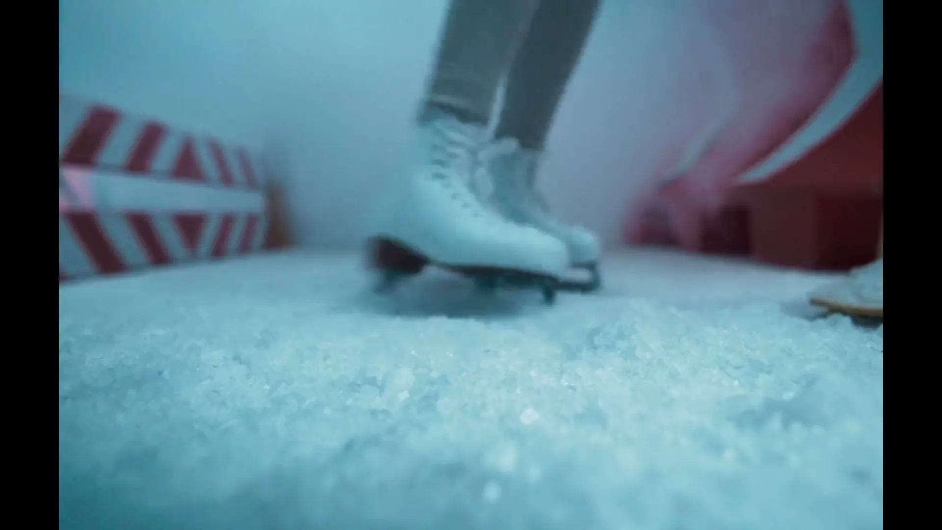 A doll's legs in white ice skates stand on a surface of crushed ice, then fall through it, in a miniature set.