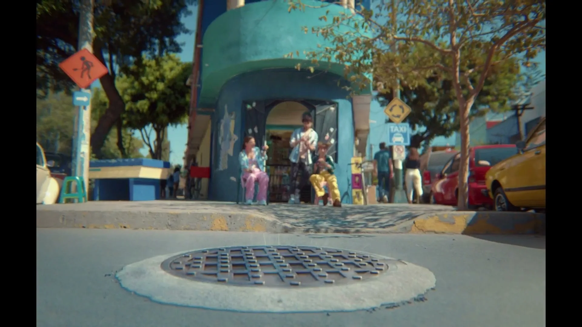 Three young people sit and stand outside a colorful building on a sunny day, as a manhole cover opens in the foreground.