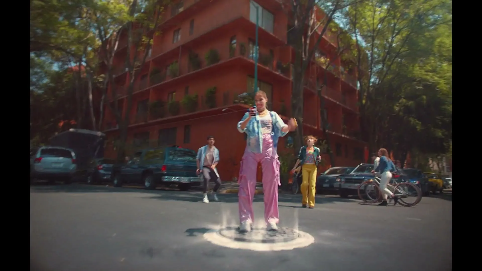 A young woman in pink pants and a blue top stands in a parking lot, surrounded by a glowing circle, as a UFO descends from above.