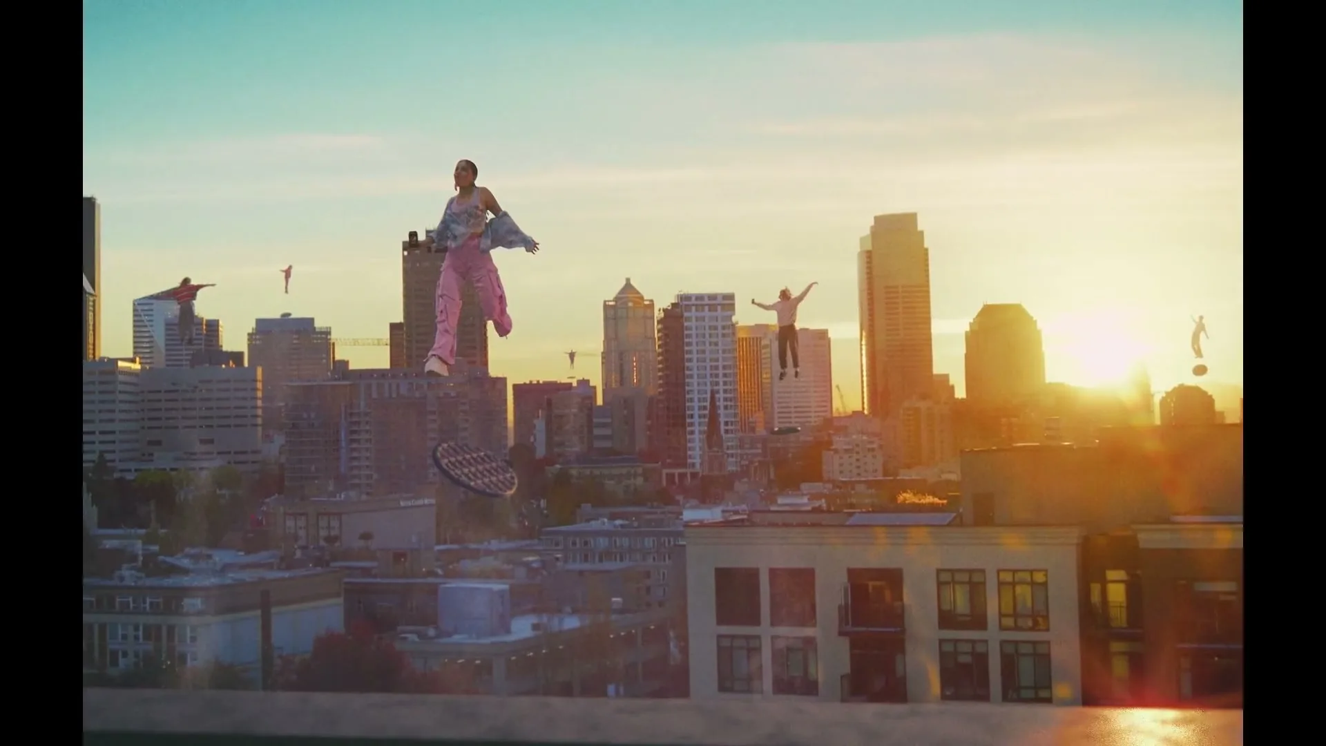 A diverse group of people float above a city skyline at sunset, with one woman in pink pants prominently in the foreground.