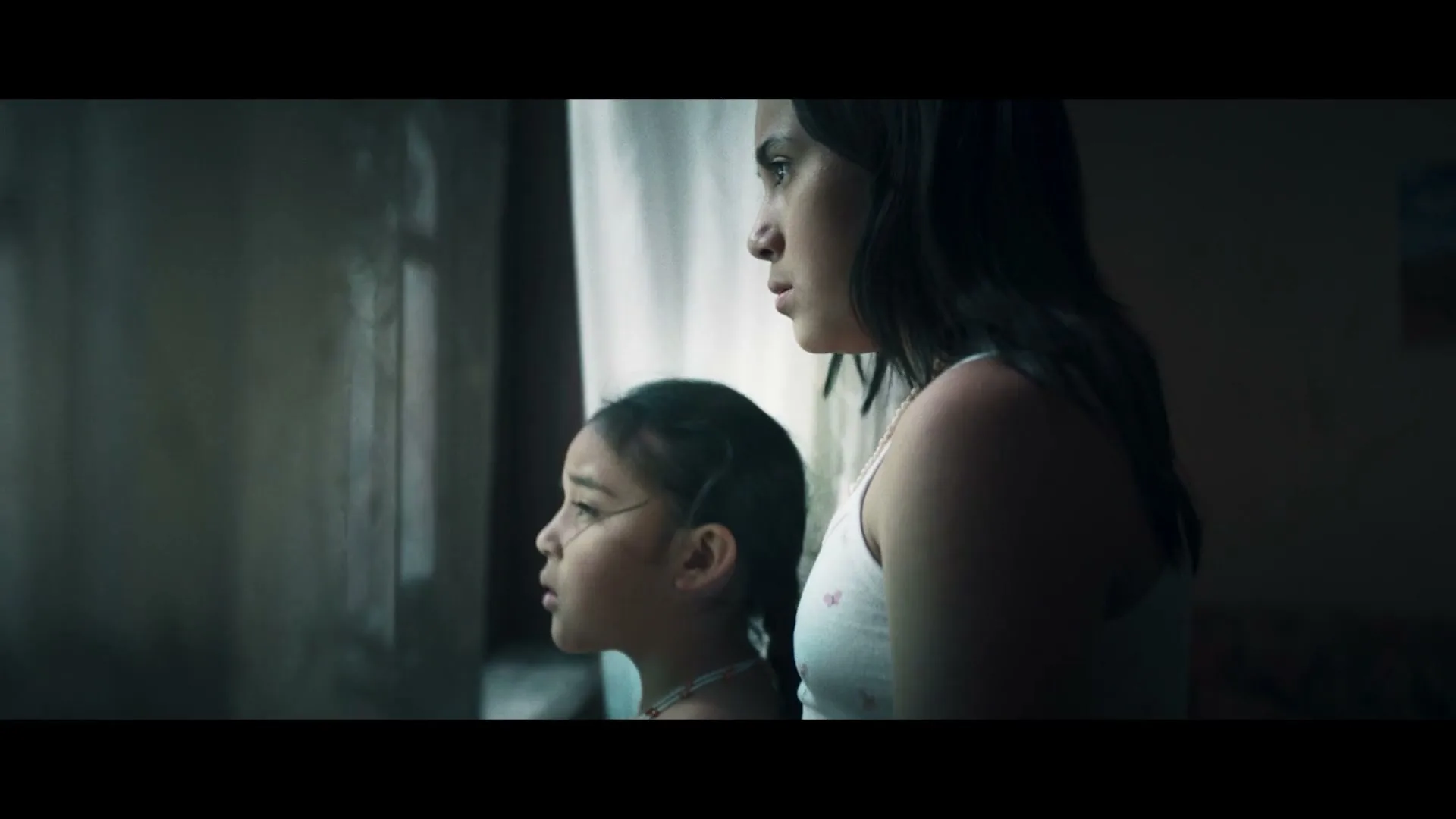 A young girl with braided hair and a woman look out a window, their faces illuminated by light filtering through sheer curtains, as a storm rages outside.