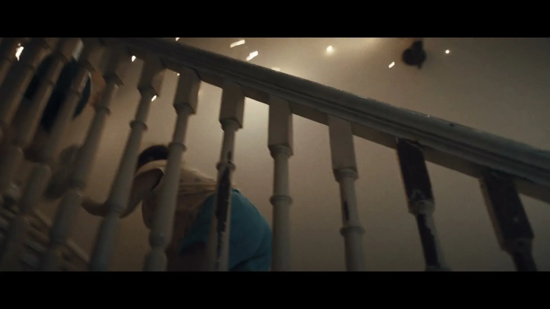 A child in blue pants and a light shirt crawls up a wooden staircase in a house, as the lighting shifts from bright to dark.
