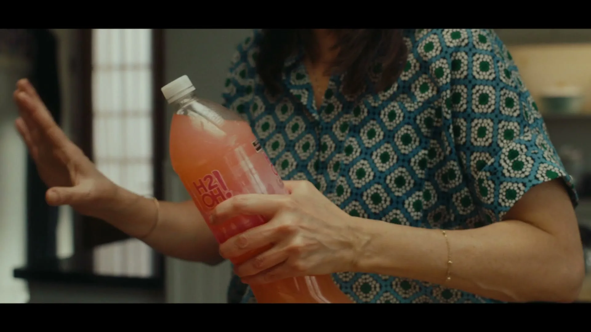 A woman in a patterned shirt holds a large bottle of pink H2oh! drink, gesticulating with her other hand in a kitchen.