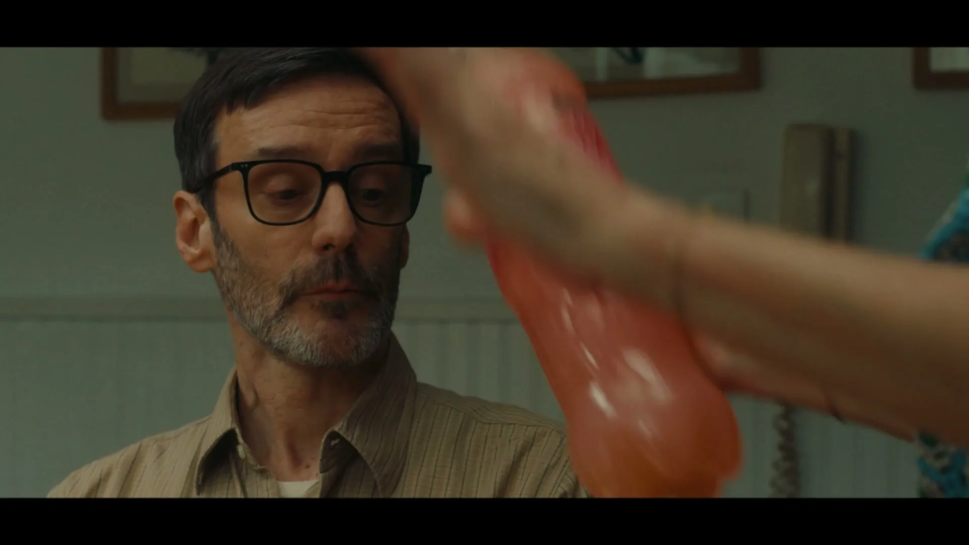 A man with glasses and a beard looks at a woman's hand holding a pink bottle, in a dimly lit room.