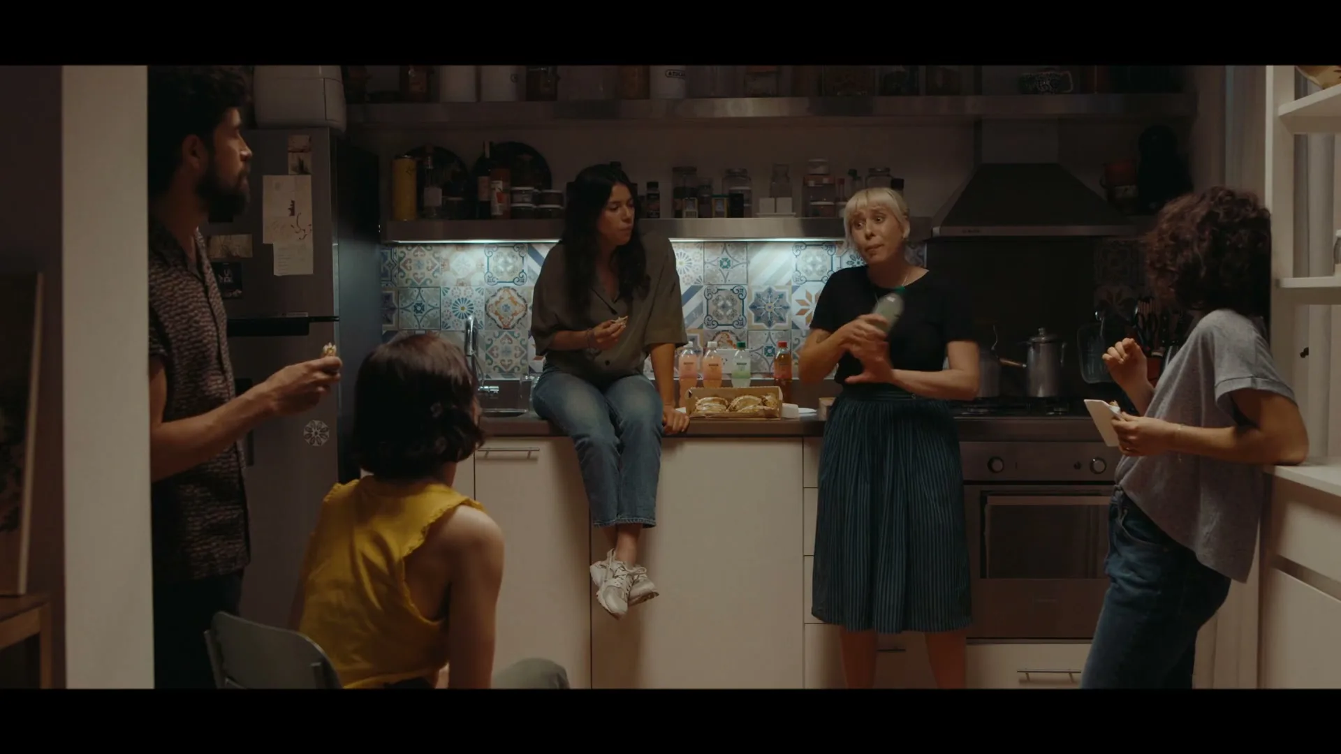 Five friends, casually dressed, gather in a cozy kitchen, eating and drinking, as one woman actively gesticulates while speaking.